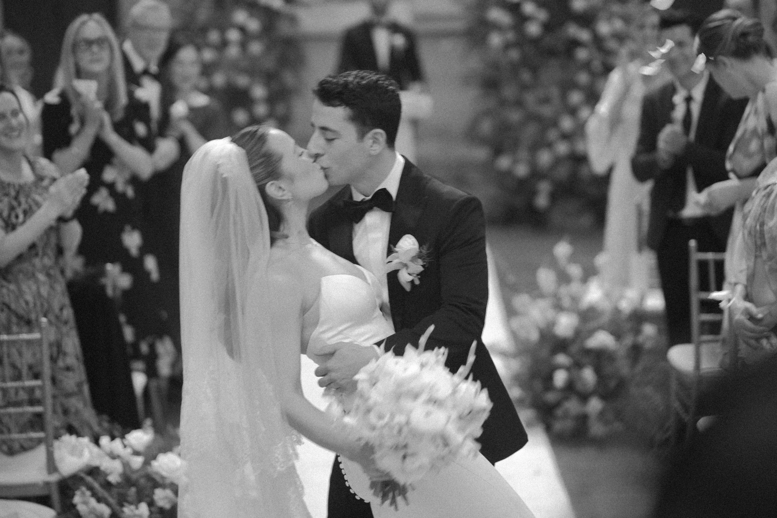 Pareja de novios besándose en su boda, rodeados de invitados en una ceremonia en interiores.