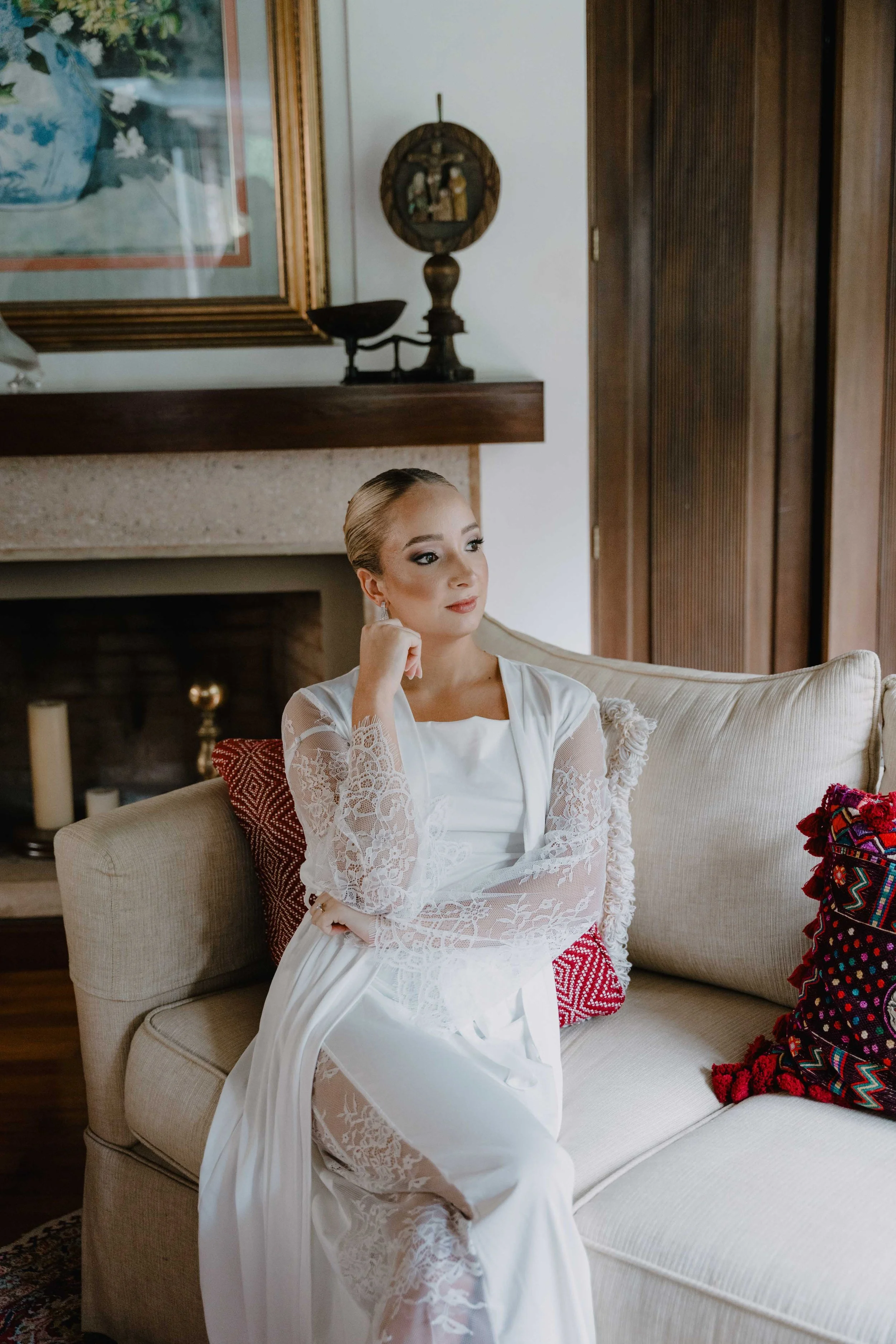 Una mujer vestida con un vestido blanco de encaje está sentada en un sofá beige en una sala con paredes blancas, madera oscura y decoración en colores cálidos.