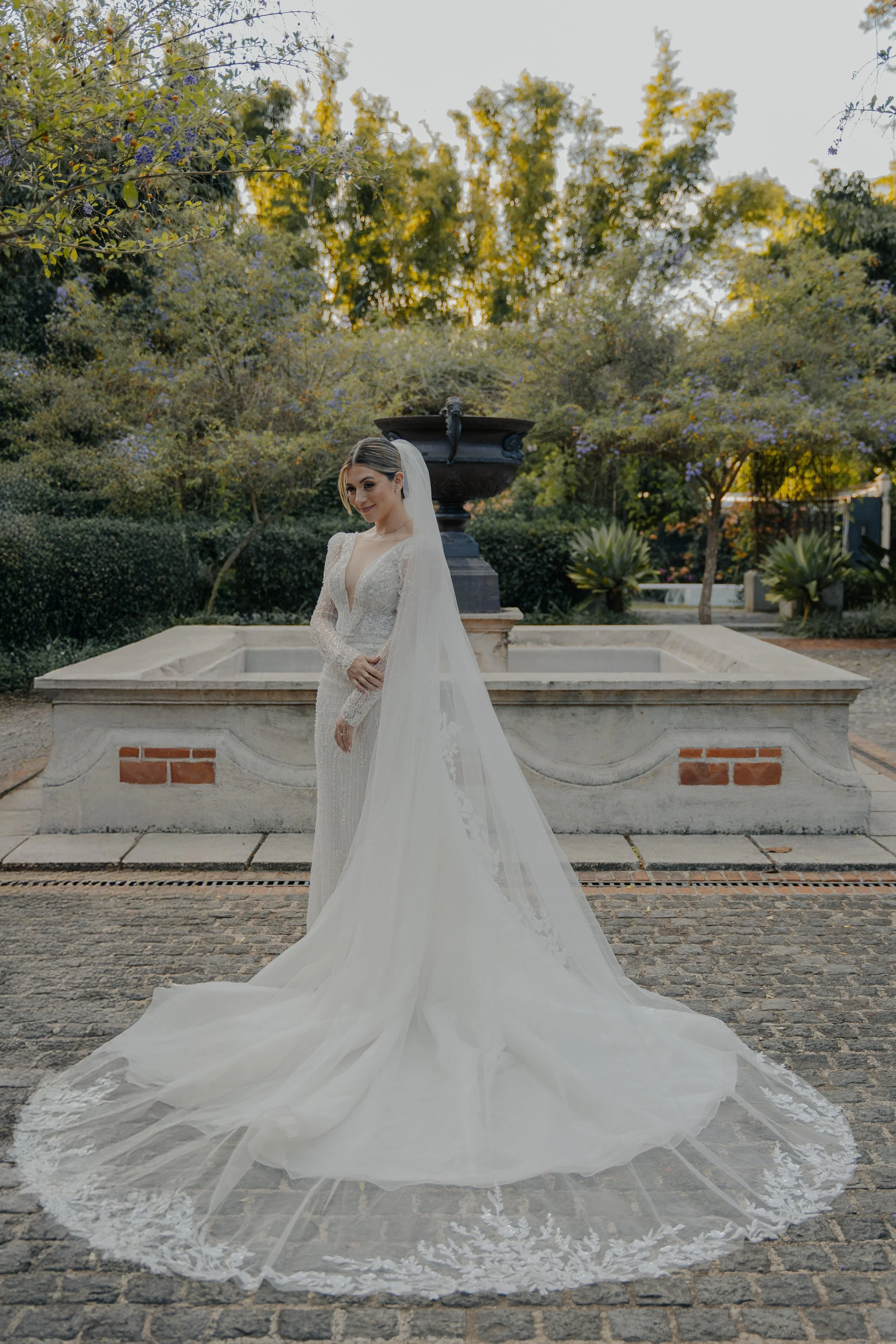 Mujer vestida de novia con vestido largo y velo en un jardín con fuente y árboles verdes.