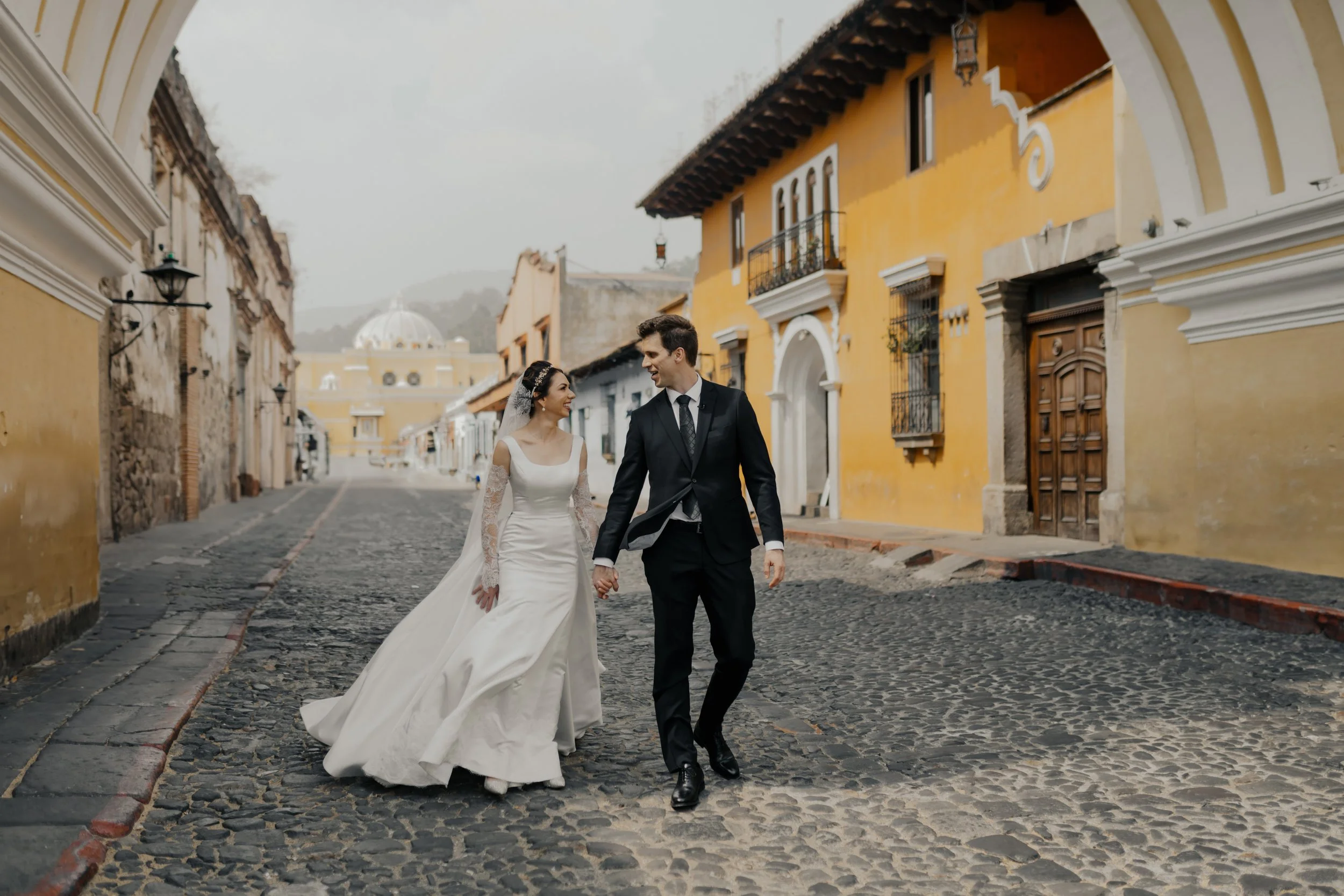Una pareja disfrazada de boda caminando por una calle empedrada en un pueblo colonial con edificios de colores amarillos y paredes blancas, en un día nublado.
