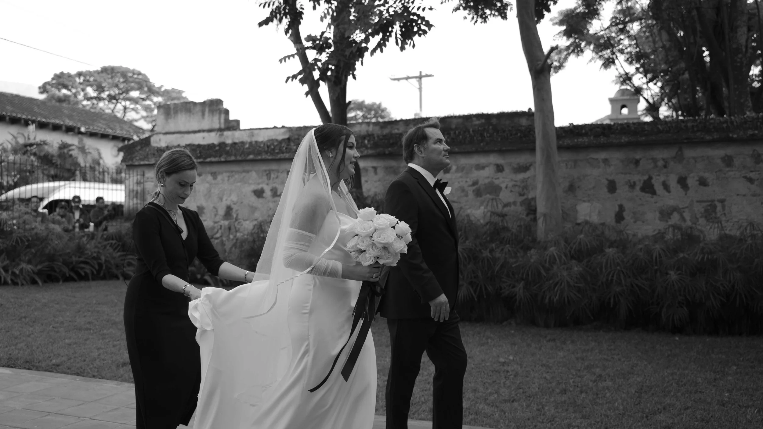 Mujer con vestido de novia sosteniendo un ramo de rosas blancas, acompañada por un hombre en traje formal, en un evento de boda exterior.