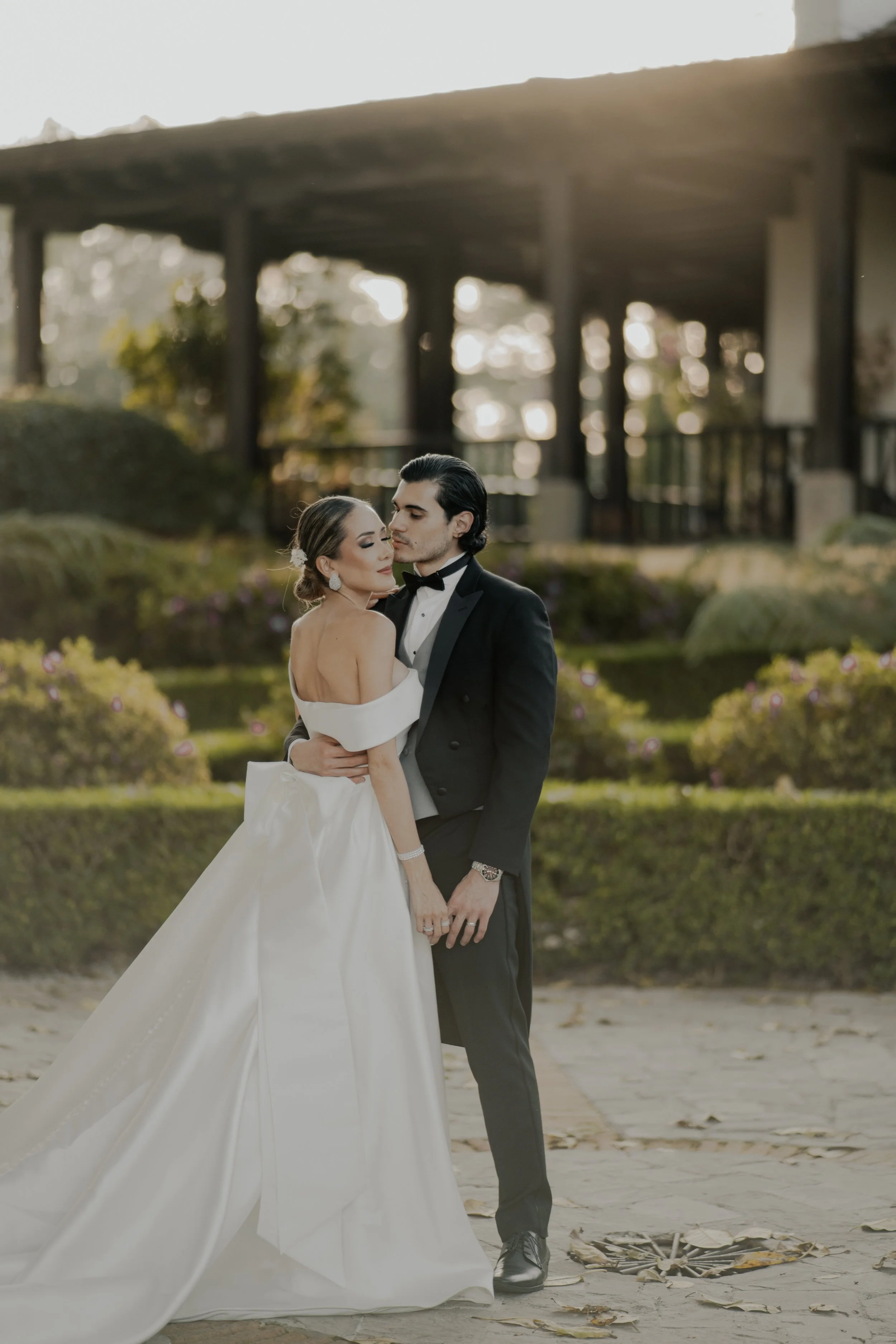 Pareja de novios en boda, el esposo con traje negro y la esposa en vestido de boda blanco, en un entorno natural con vegetación y estructura de madera en el fondo.