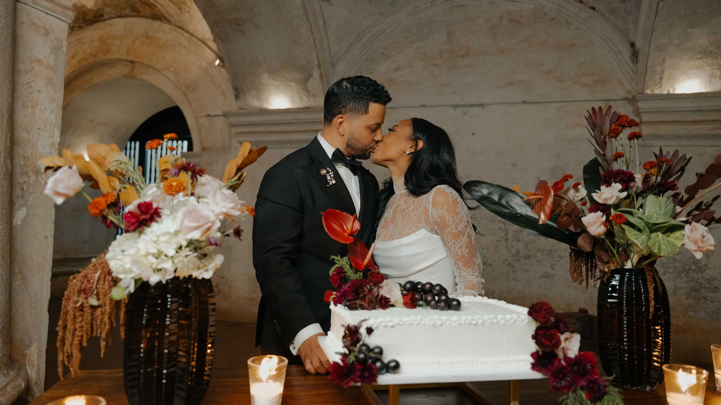 Una pareja de recién casados besándose frente a un pastel de bodas decorado con frutas y flores en una ceremonia en interiores.