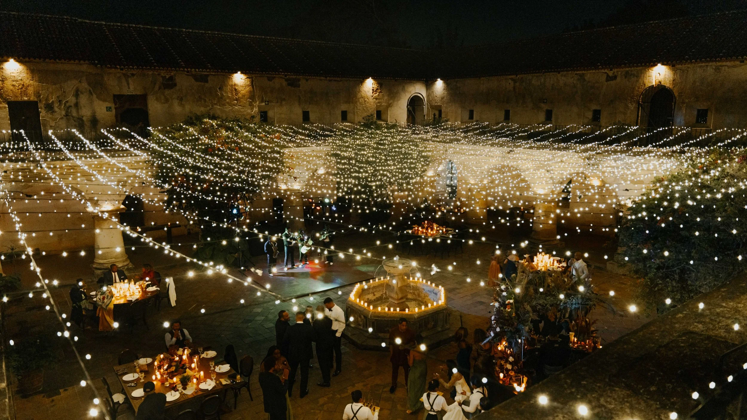 Celebración al aire libre en la noche con iluminación de cadenas de luces, mesas decoradas, personas bailando y conversando en un espacio con fuente en el centro y paredes antiguas.