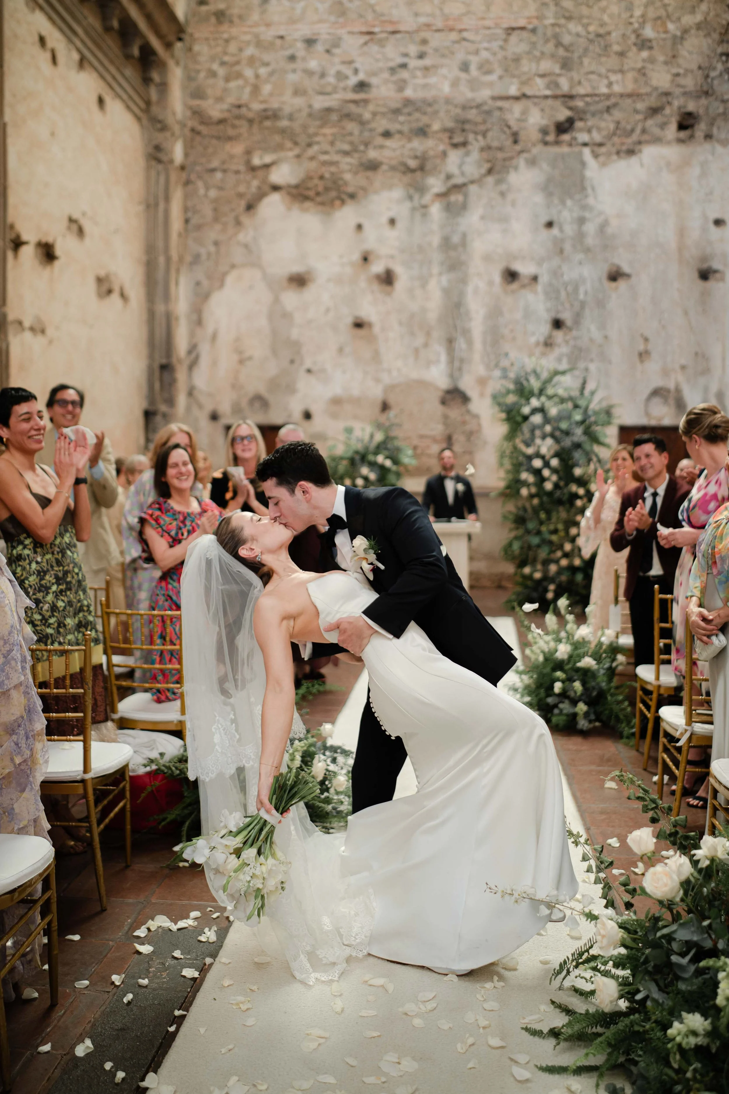 Pareja de novios en su boda, el novio la dobla y la besa en la ceremonia, rodeados de invitados en un lugar con paredes de piedra y decoraciones florales blancas.