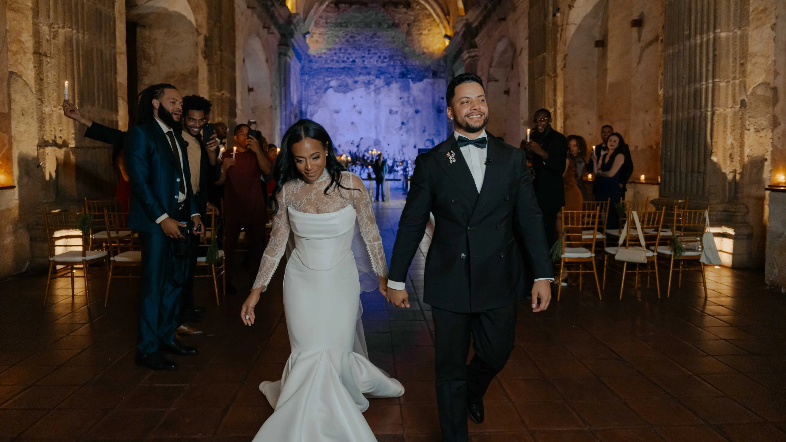 Pareja de recién casados caminando por una iglesia antigua decorada con flores, mientras sus invitados los observan y toman fotos, en una boda elegante y emotiva.