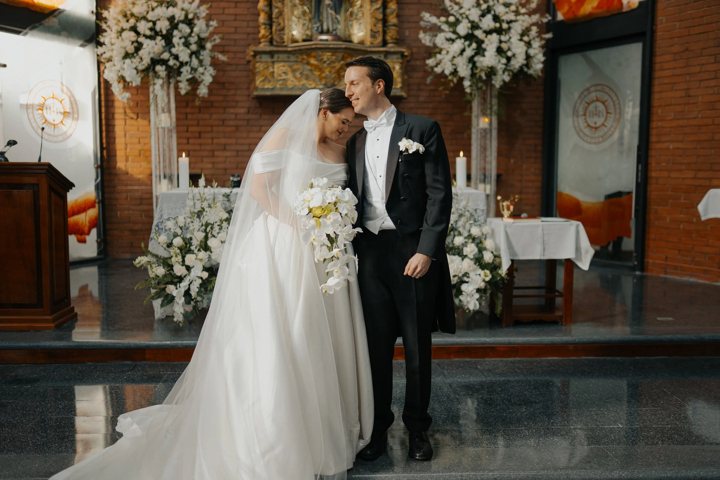 Pareja de recién casados en la ceremonia religiosa, la novia con vestido blanco y velo, el novio con traje de etiqueta negro, en un altar con flores blancas y velas.