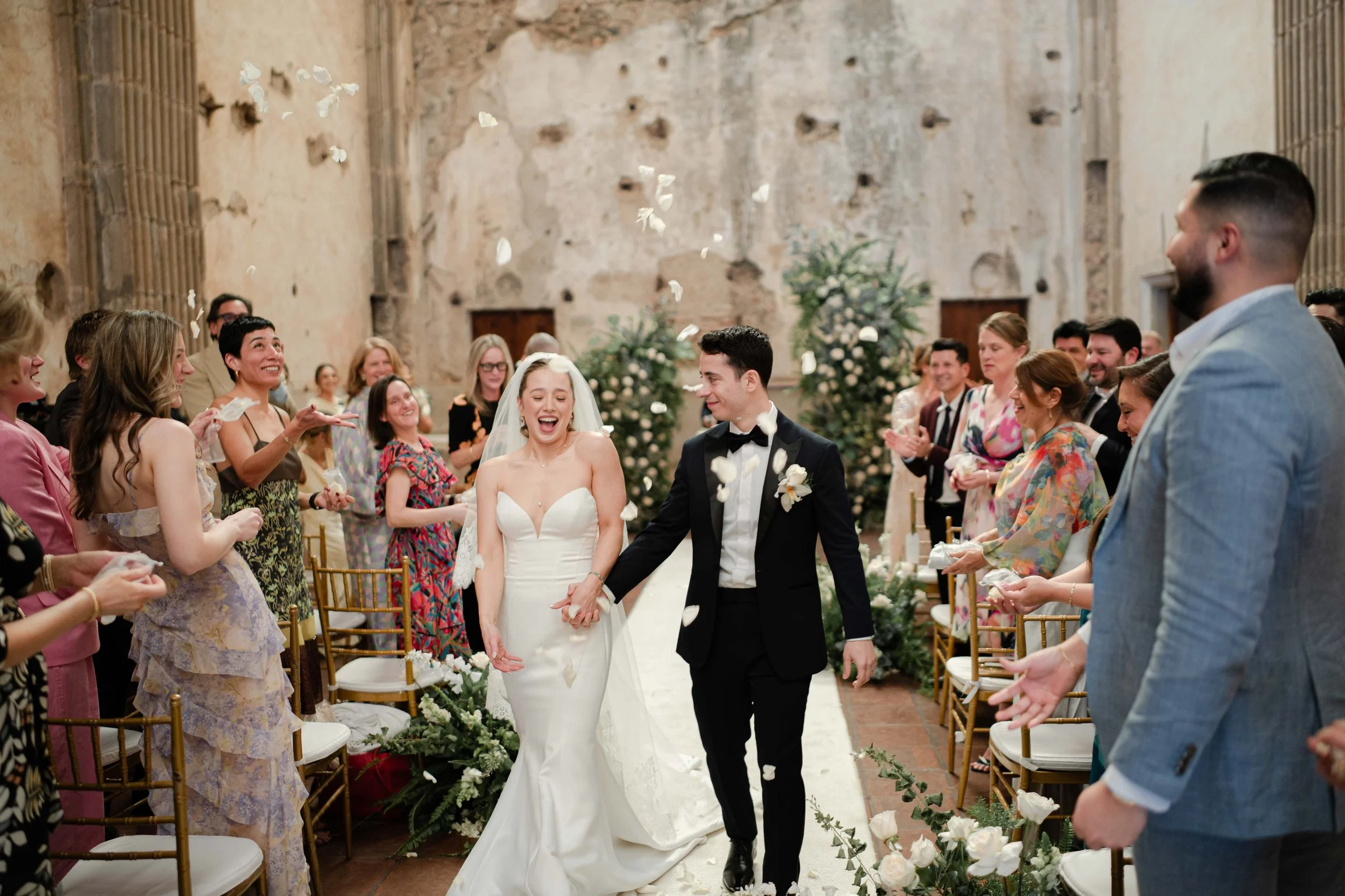 Una pareja de casados caminando por una alfombra decorada, rodeados de amigos y familiares, lanzando pétalos de rosa en una boda en un lugar antiguo con paredes de piedra y decoraciones florales.