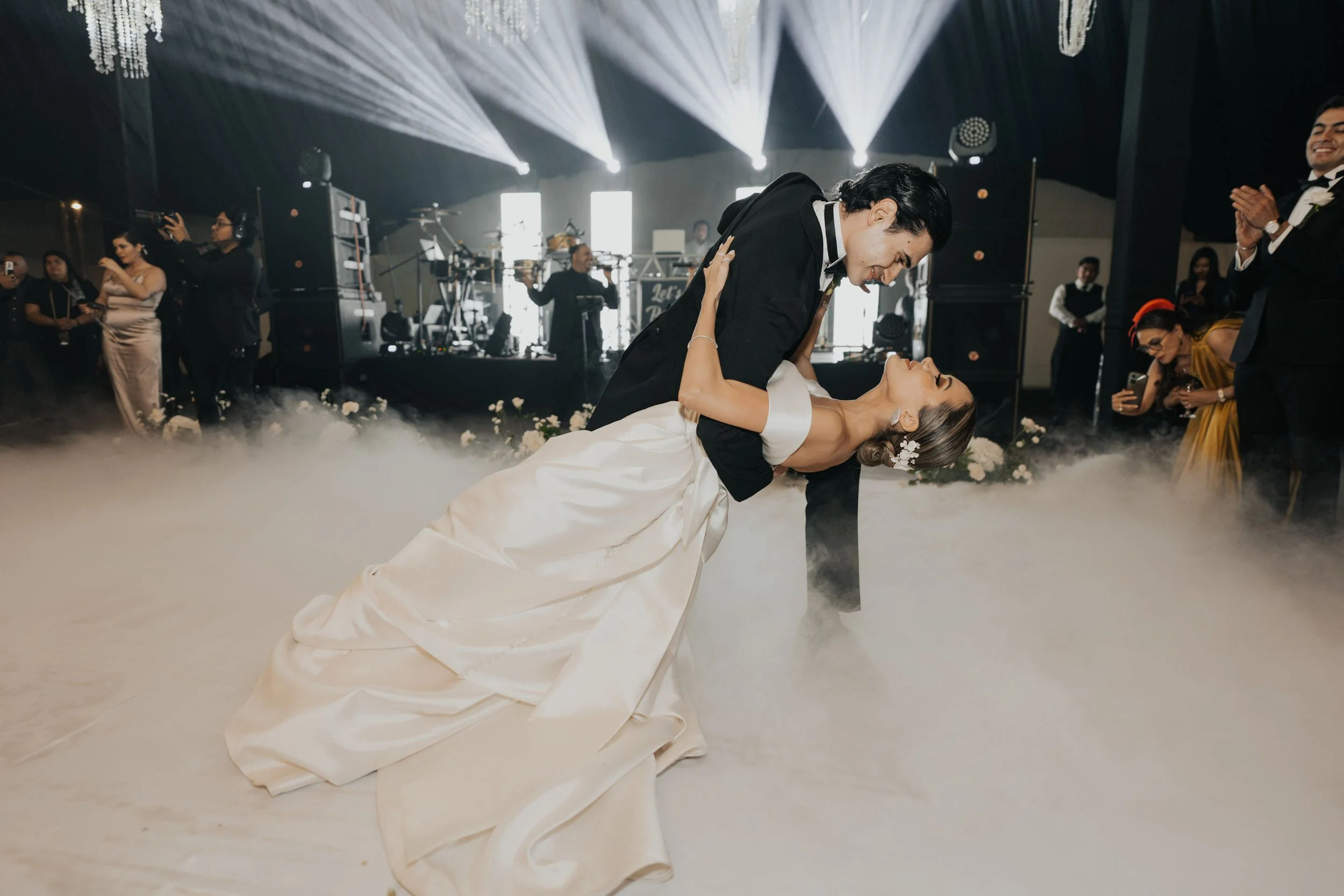 Pareja de recién casados bailando en su boda, rodeados de invitados y banda en vivo en un ambiente elegante con humo en el suelo.