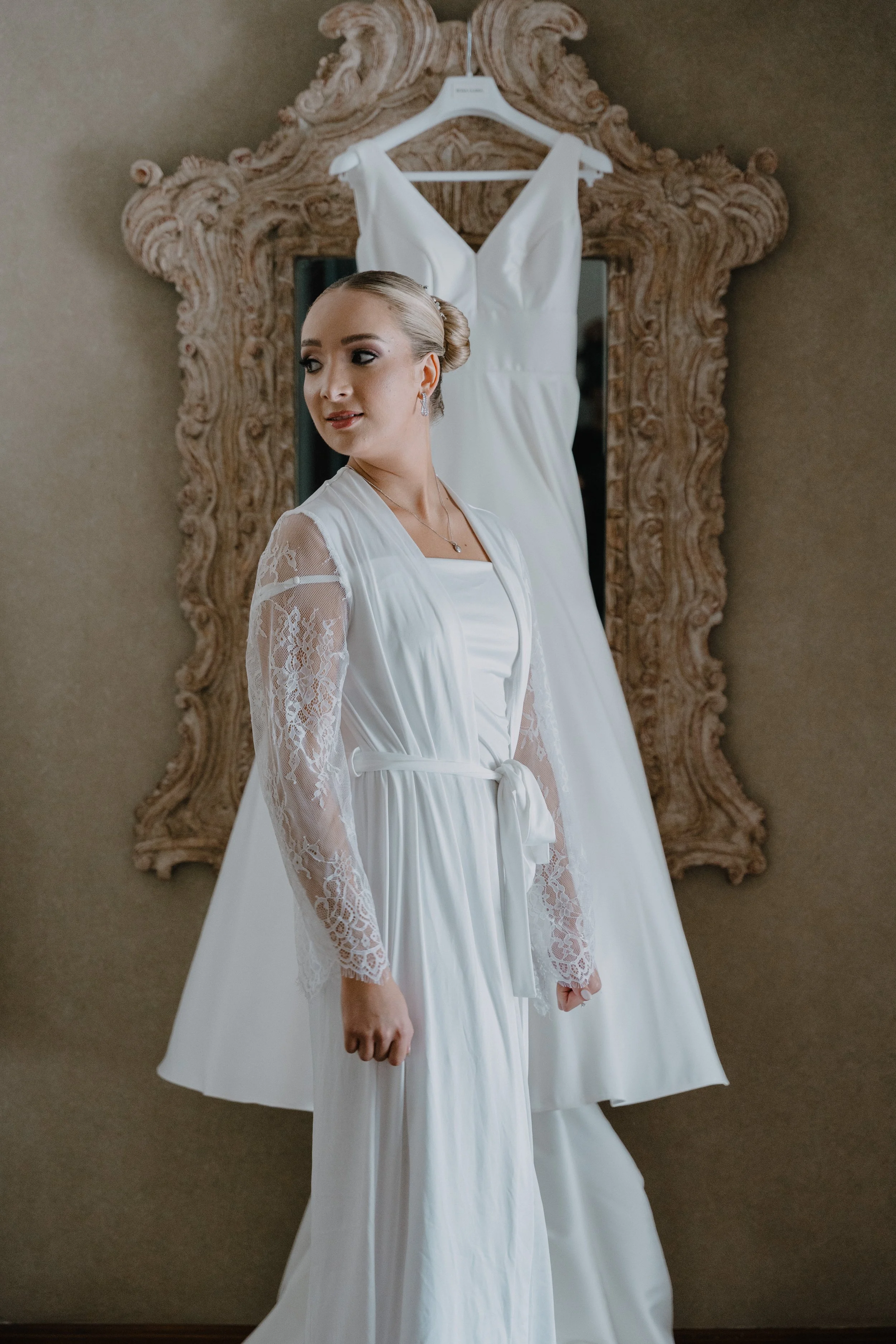 Mujer en vestido blanco de novia en interior con un espejo con marco ornamentado y vestido de novia suspendido detrás de ella.