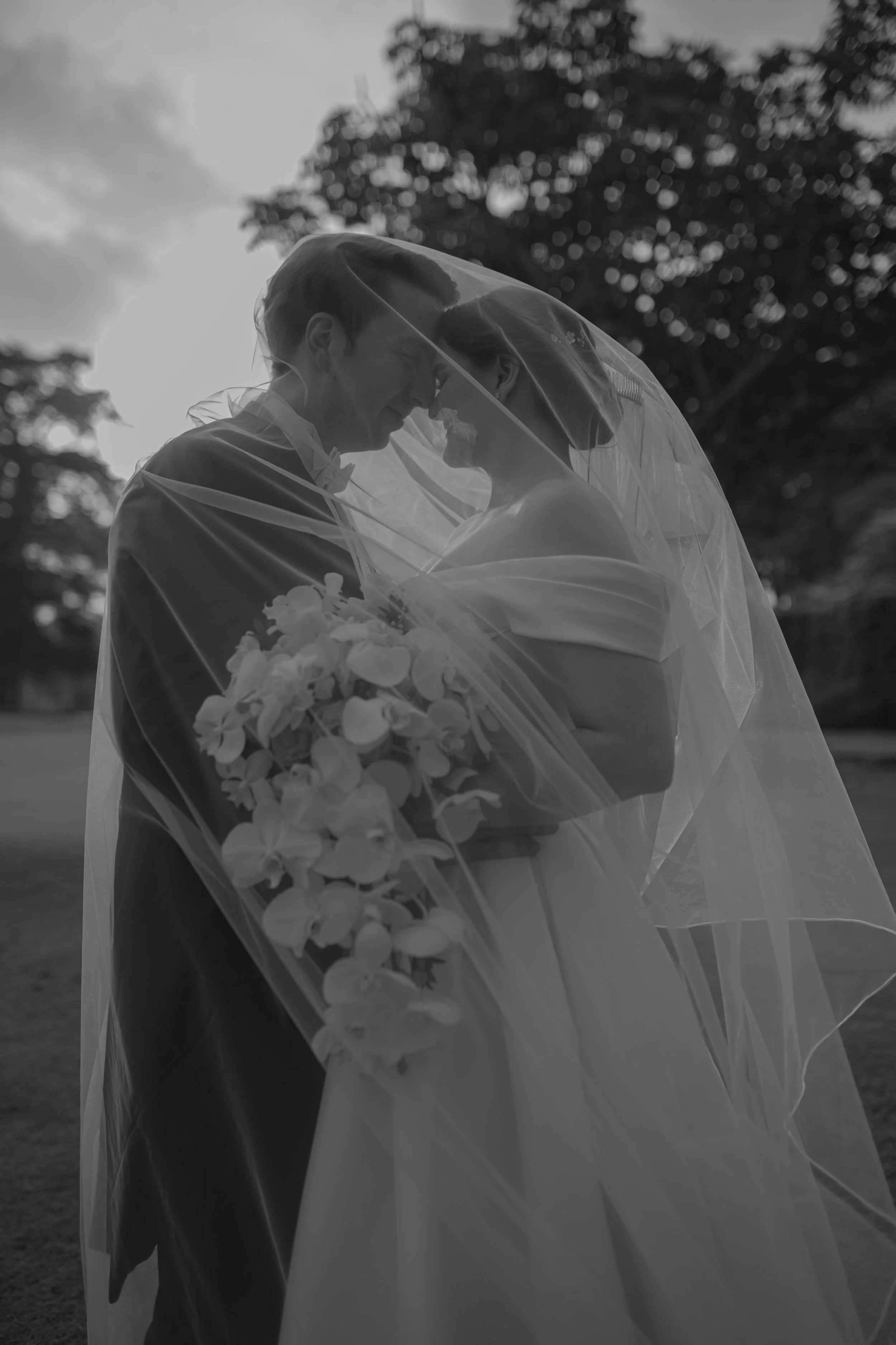 Novios de boda, con una boda en exteriores, compartiendo un momento íntimo bajo un velo, la novia sostiene un ramo de flores.