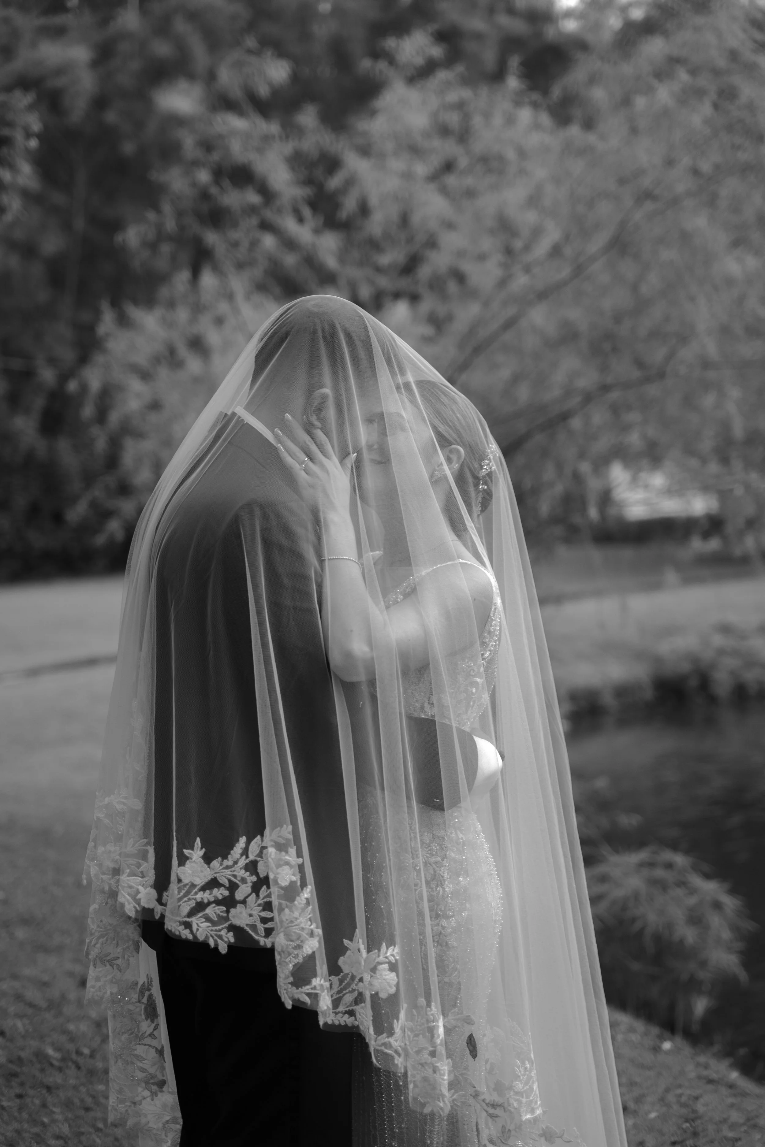 Una pareja de novios en un beso, con un velo de novia cubriéndolos, en un entorno natural al aire libre, en blanco y negro.
