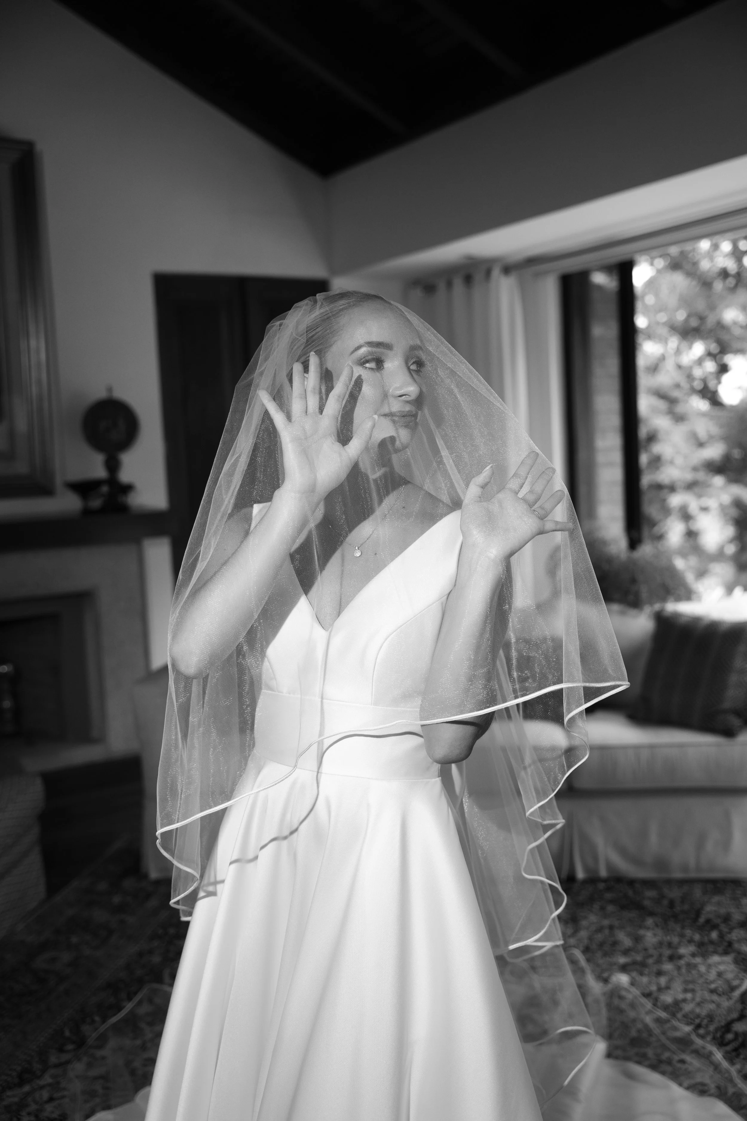 Mujer con vestido de novia y velo negro y blanco, posando detrás de una cortina de cristal en una habitación con ventana grande que muestra árboles en el exterior, en una fotografía en blanco y negro.