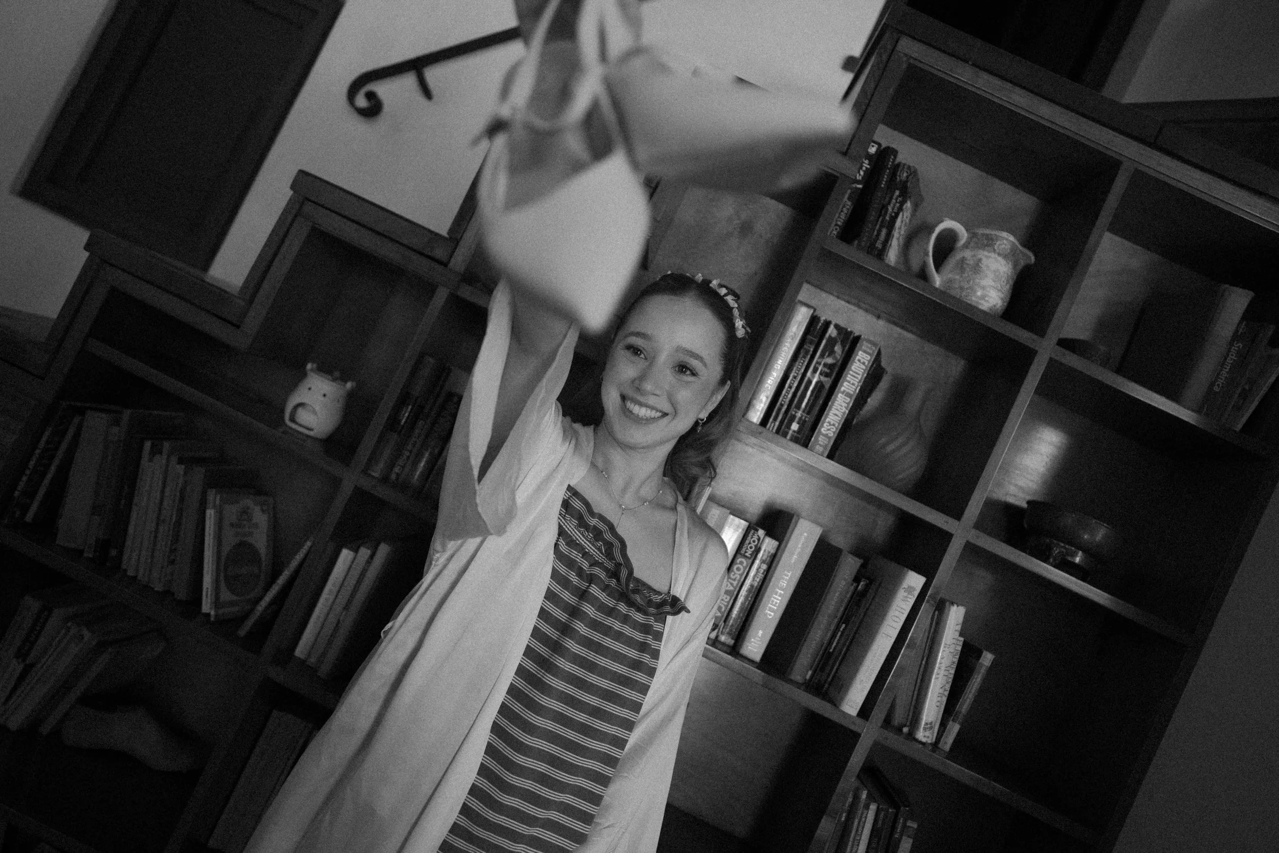 Mujer joven celebrando con una sonrisa, sosteniendo un diploma o certificado en una mano, en una sala con estanterías llenas de libros y objetos decorativos, en blanco y negro.