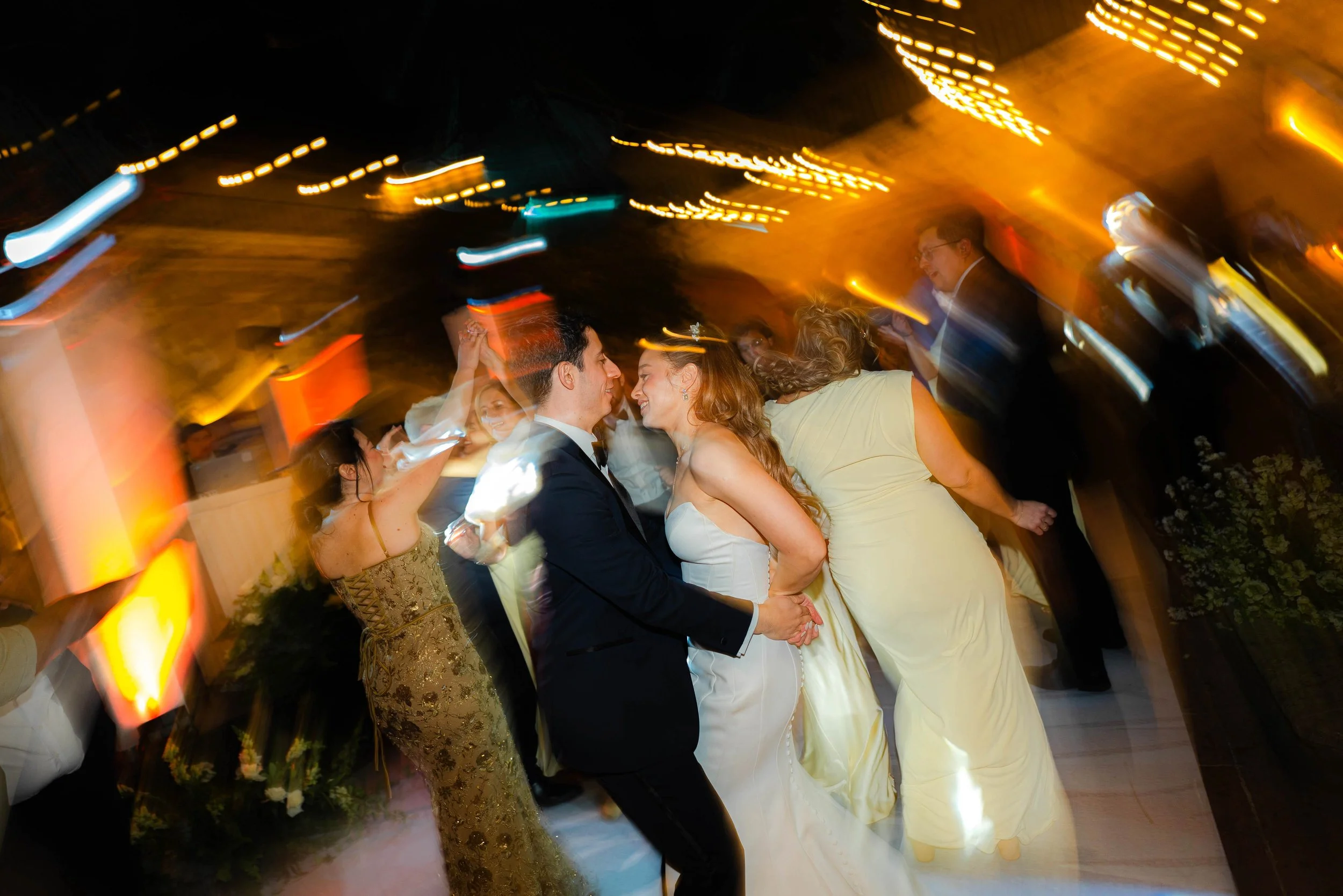 Pareja de novios bailando en una fiesta con luces de colores y otros invitados en el fondo.