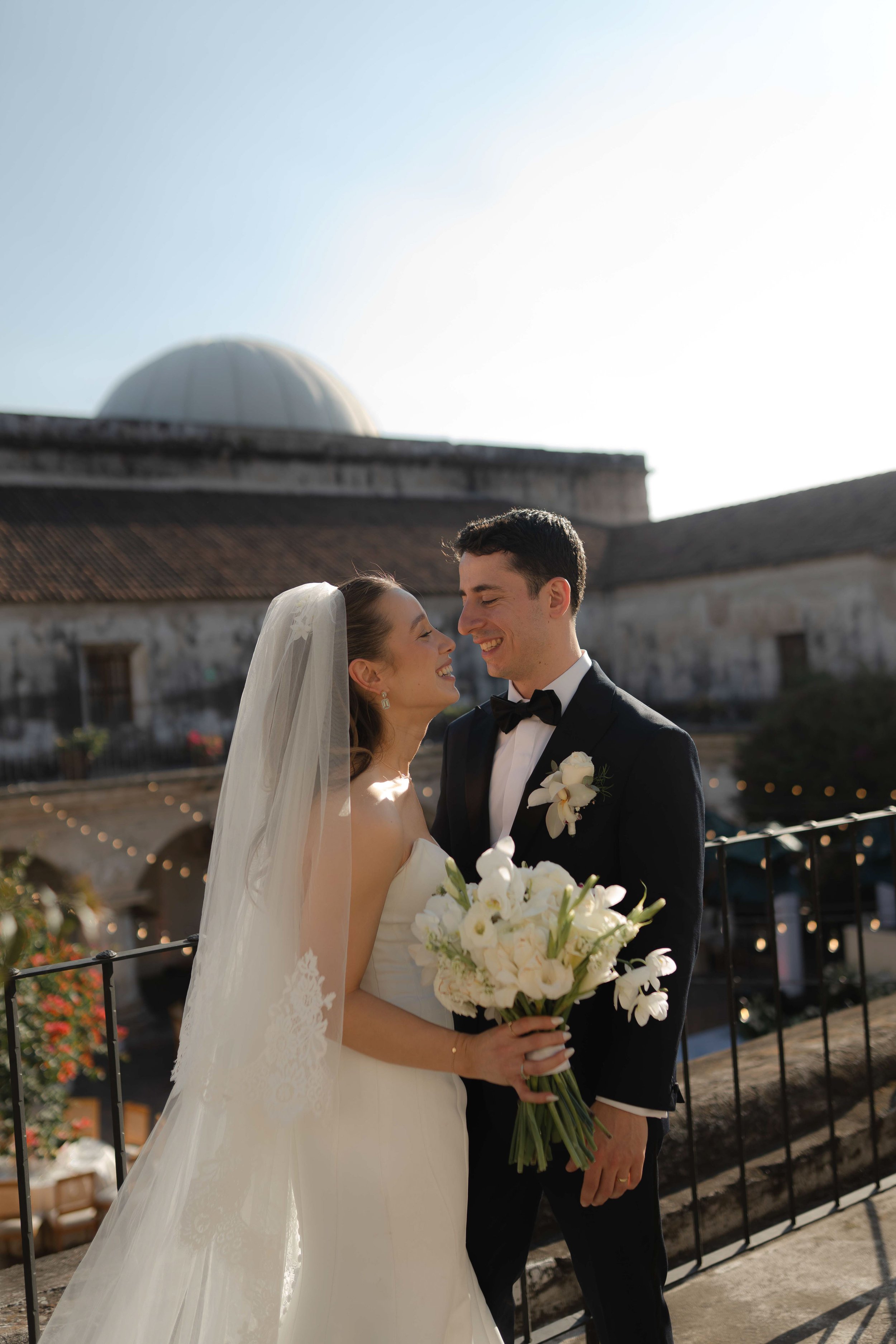 Una pareja de recién casados sonriendo y mirándose en una boda al aire libre, con un edificio antiguo y un domo en el fondo, durante el atardecer.