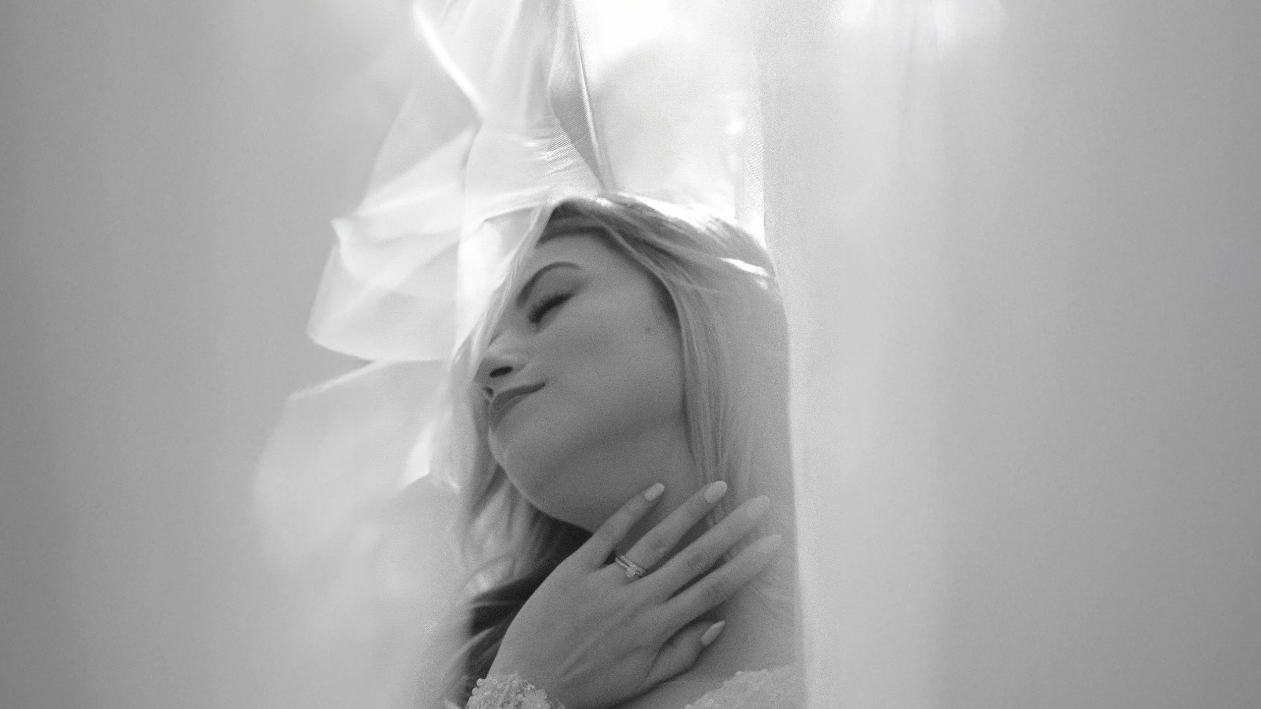 Retrato en blanco y negro de una mujer con los ojos cerrados tocándose el cuello, rodeada de telas traslúcidas y ligeras, con expresión tranquila y serena.