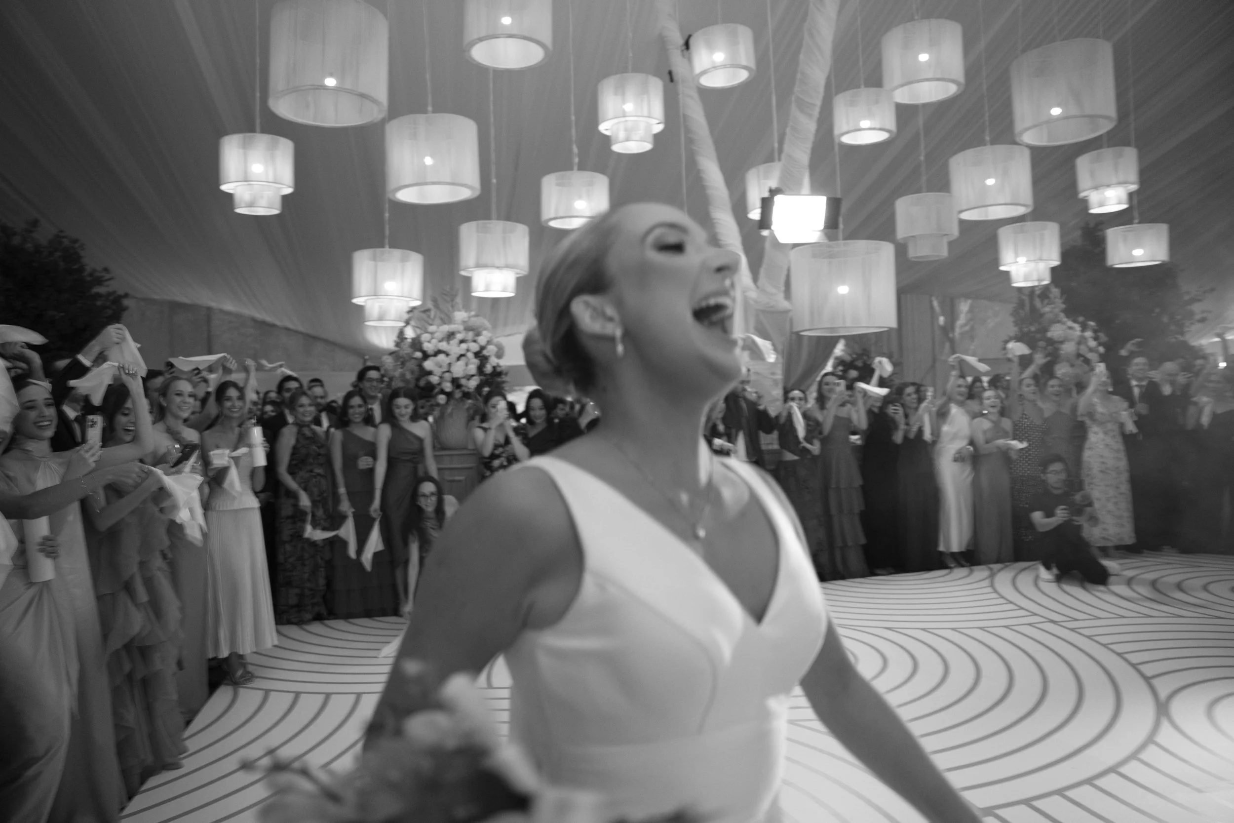 Mujer sonriendo en un evento con muchas personas, decoraciones y lámparas colgantes.