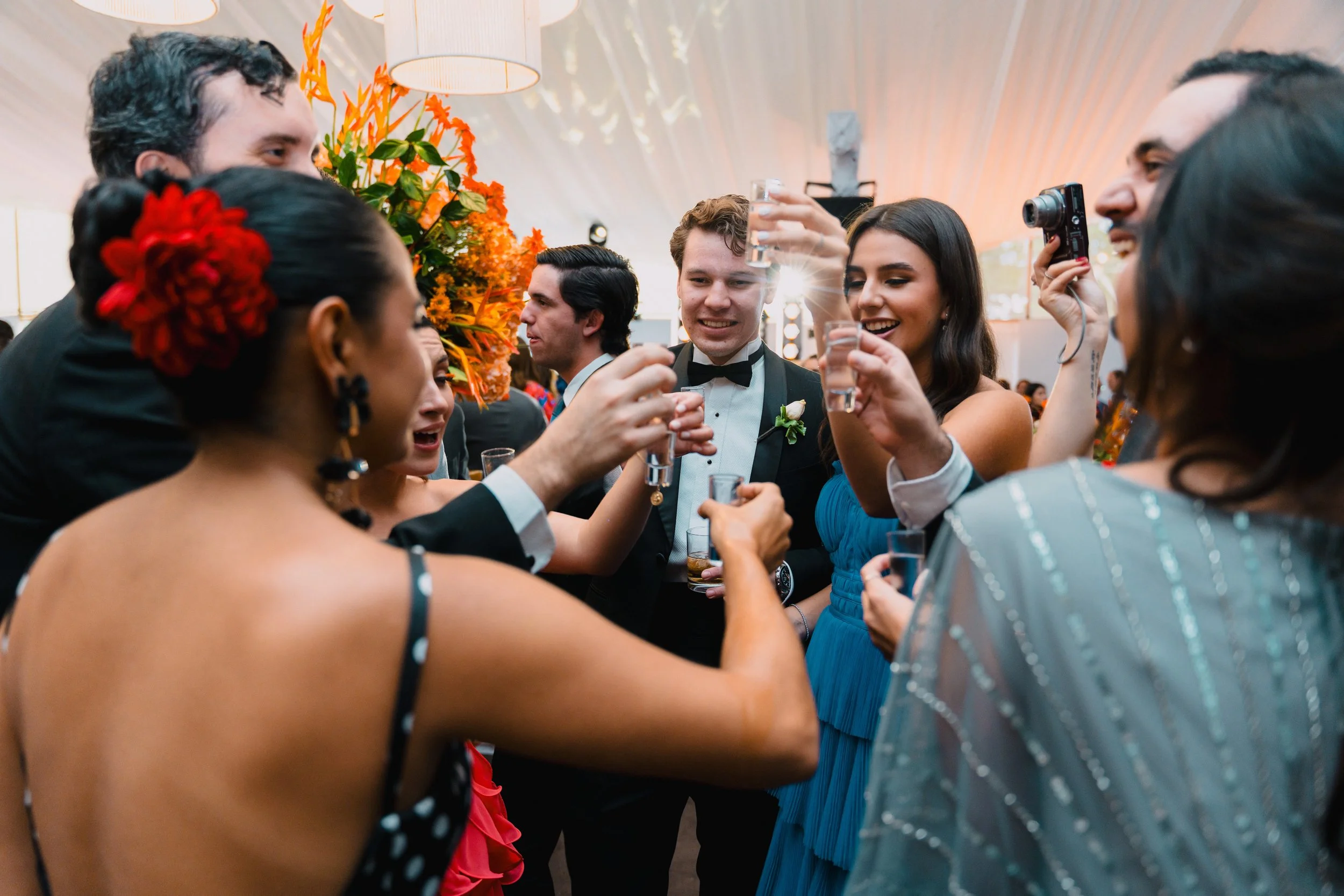 Grupo de personas en un evento social celebrando con copas en mano y sonrientes, con decoraciones florales y ambiente festivo.