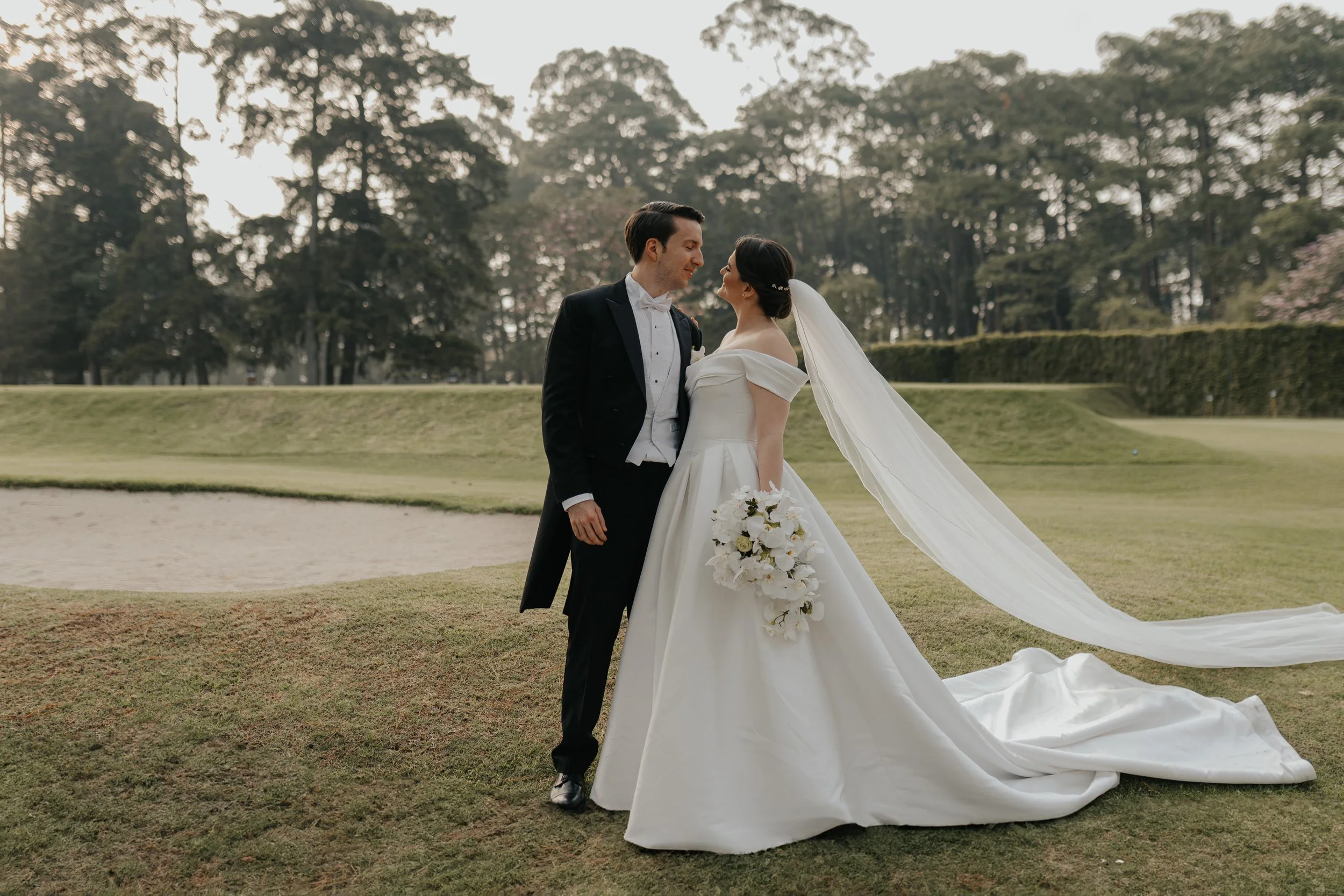 Pareja en su boda en un campo con árboles en el fondo, la novia con vestido largo y velo, el novio con traje formal, acariciándose y sonriendo.