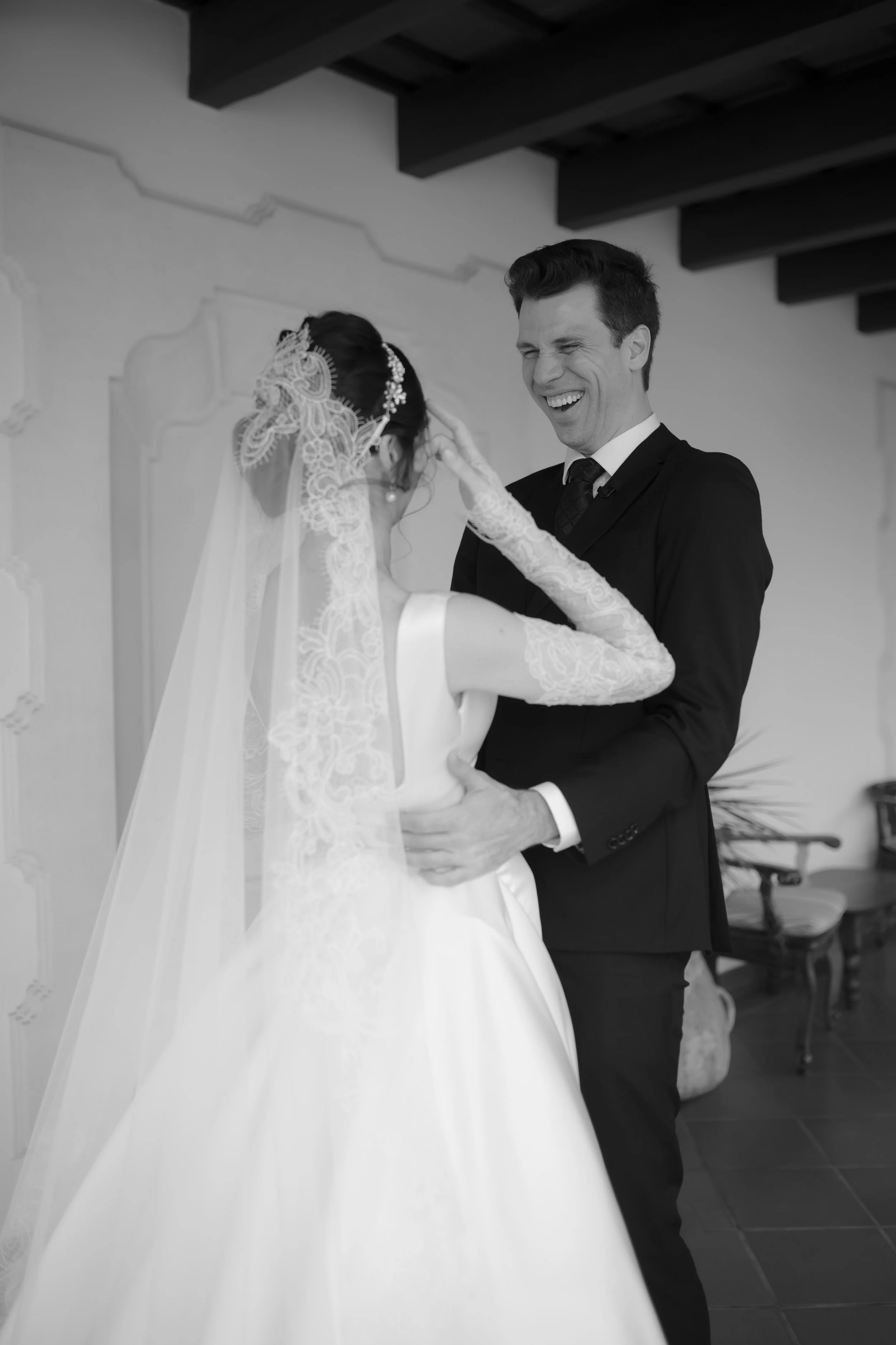 Una pareja de novios riendo, la novia con vestido de novia y velo, el novio con traje oscuro, en una celebración de boda en interiores.