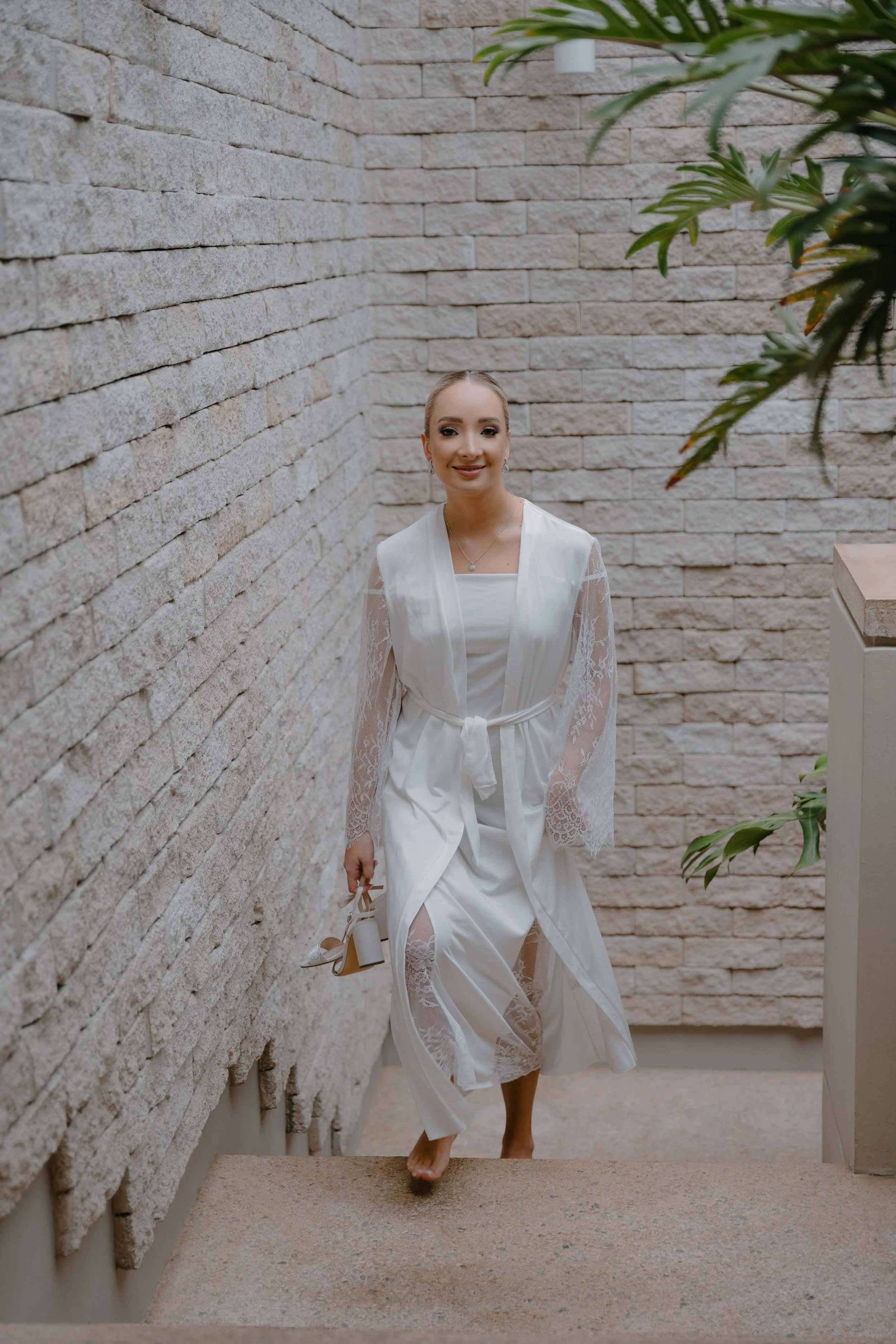 Mujer en vestido blanco y ropa de seda, bajando unas escaleras en un espacio con paredes de piedra beige, sosteniendo un par de zapatos de tacón y sonriendo.