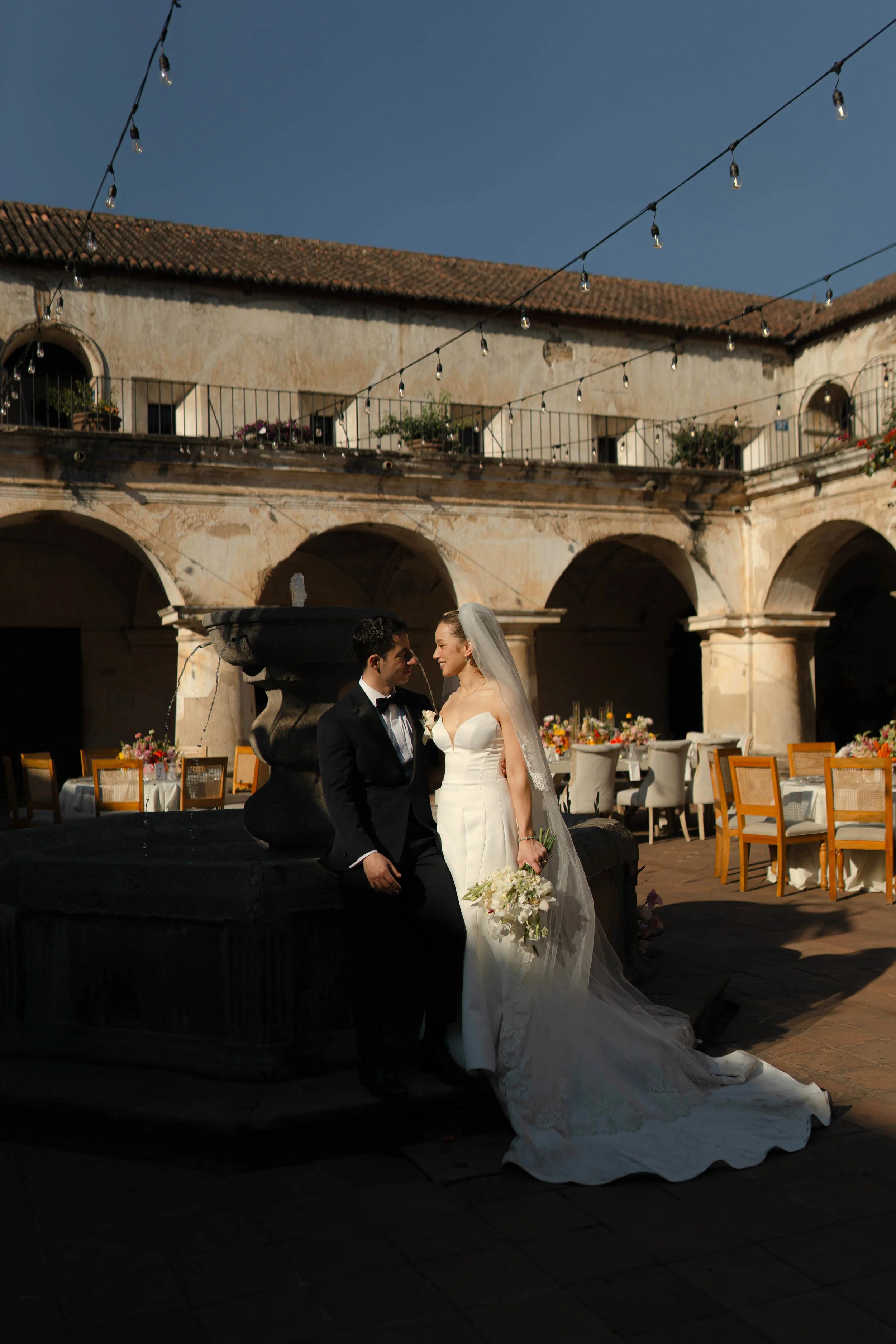 Una pareja de recién casados en un patio con una fuente en el centro, mesas decoradas con flores, y un cielo despejado.