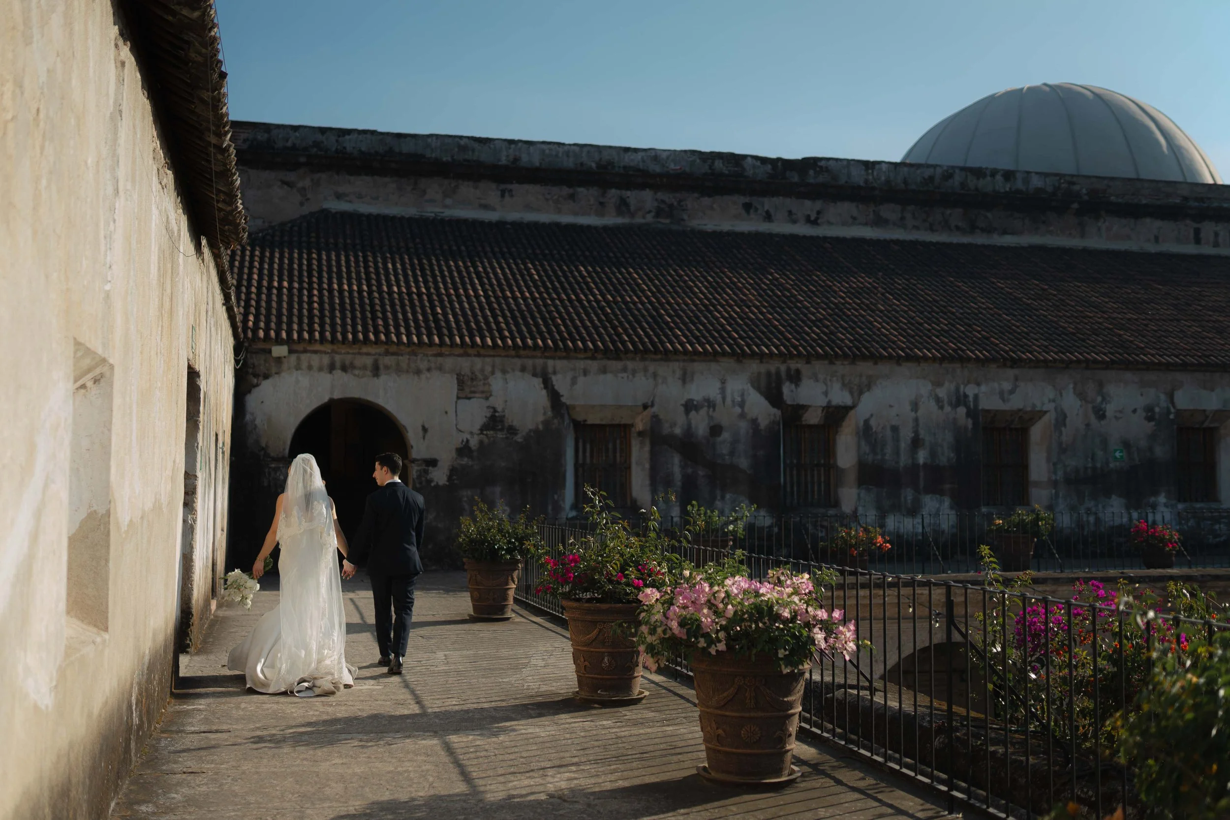 Una pareja de novios caminando juntos por un callejón empedrado con paredes de piedra y macetas con flores coloridas, con un cielo despejado y un edificio antiguo en el fondo.