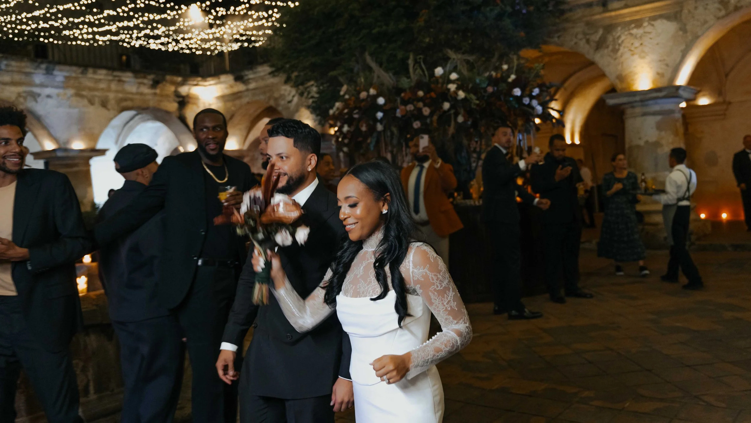 Una pareja en vestido de novia y traje de novio bailando y sonriendo en una celebración con asistentes en un ambiente elegante y decorado con flores y luces cálidas.