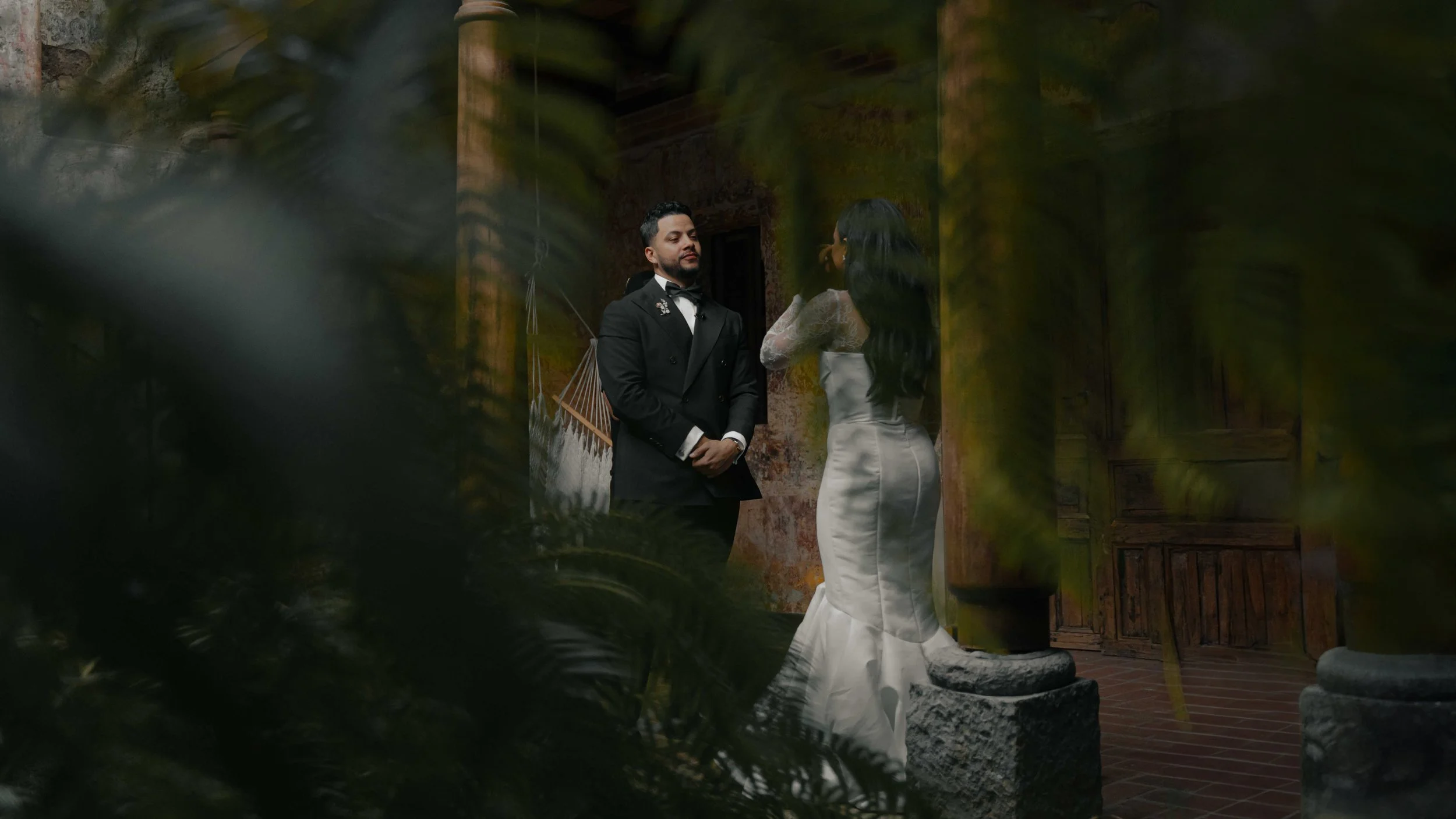 Pareja en vestimenta formal en un lugar antiguo con paredes de ladrillo y columnas de piedra, parcialmente cubiertos por vegetación.