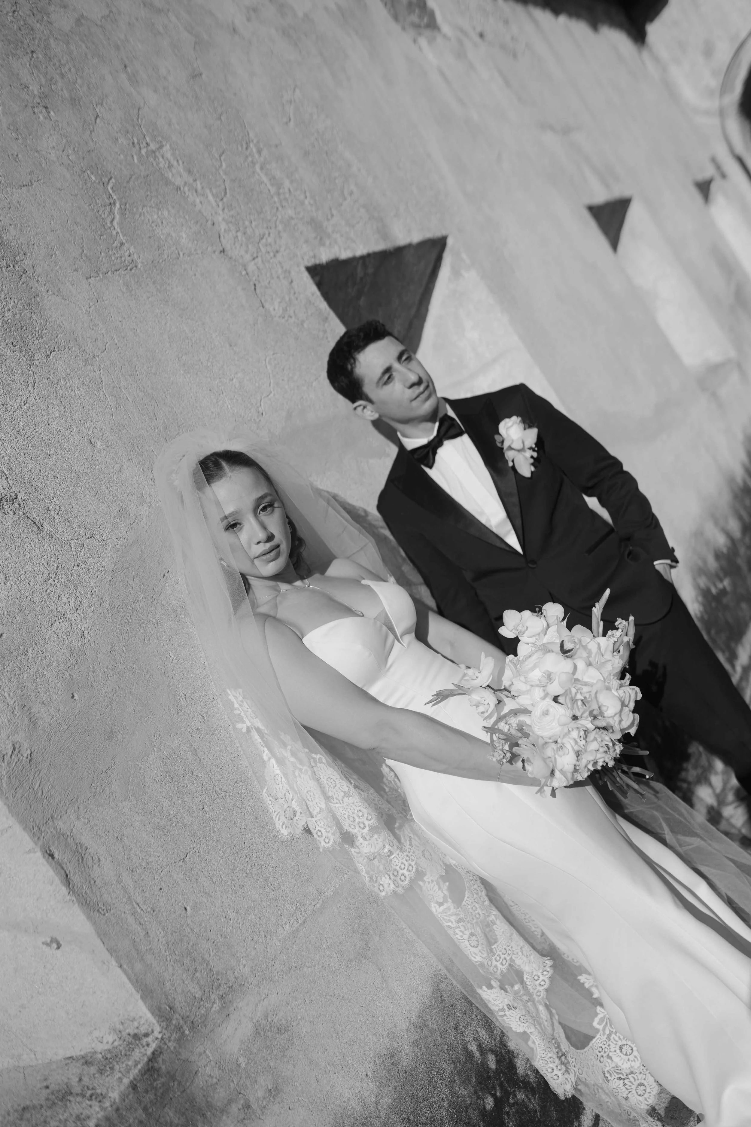 Una pareja de novios de espaldas a la pared, en la boda. La mujer lleva un vestido de novia y un velo, sostiene un ramo de flores blancas, y el hombre lleva un traje negro con pajarita y una flor en la solapa.