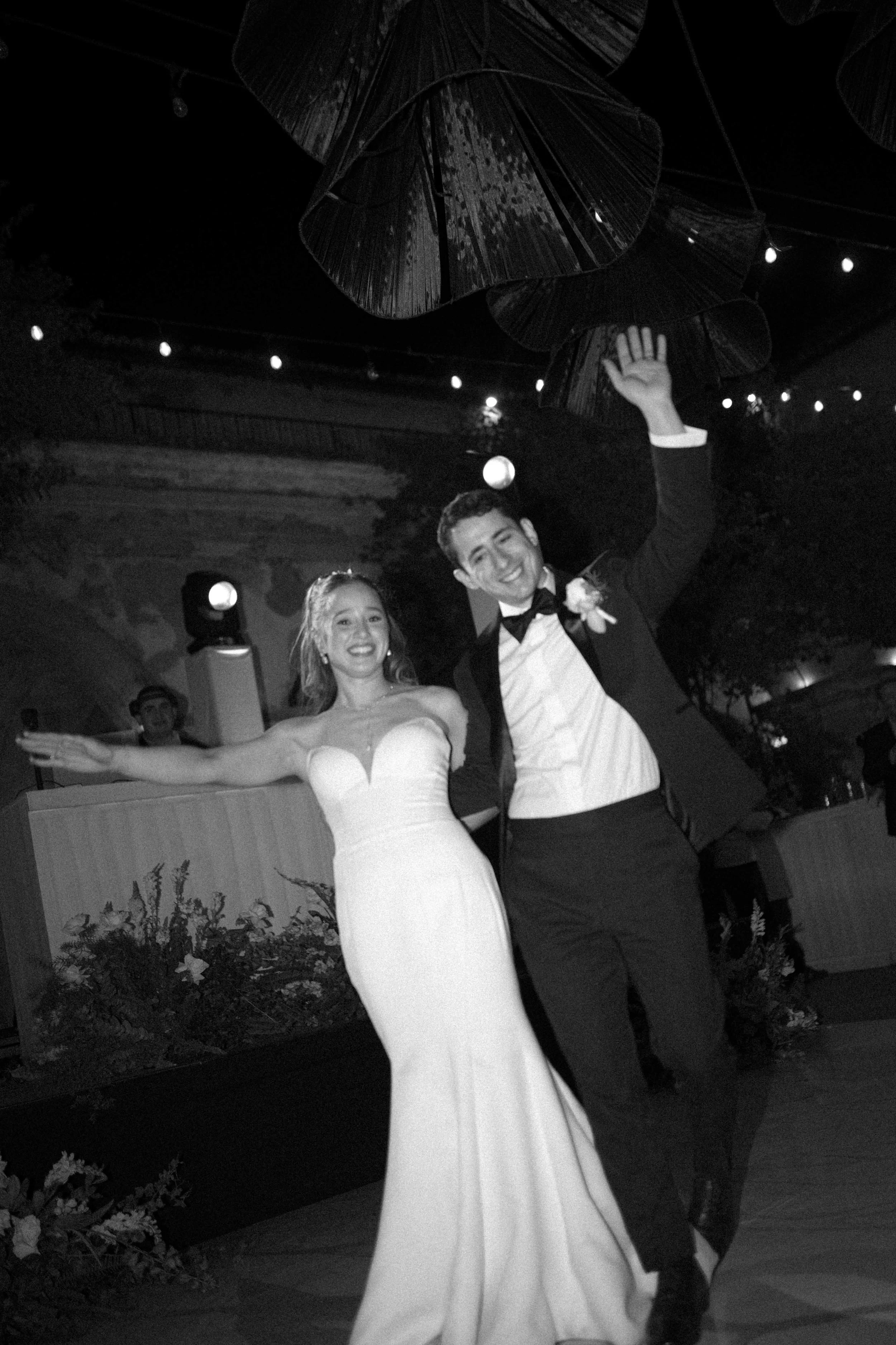 Pareja bailando en una boda, la novia lleva vestido blanco largo y el novio lleva traje formal con pajarita, ambos sonrientes. La escena es en un lugar decorado con luces y plantas.
