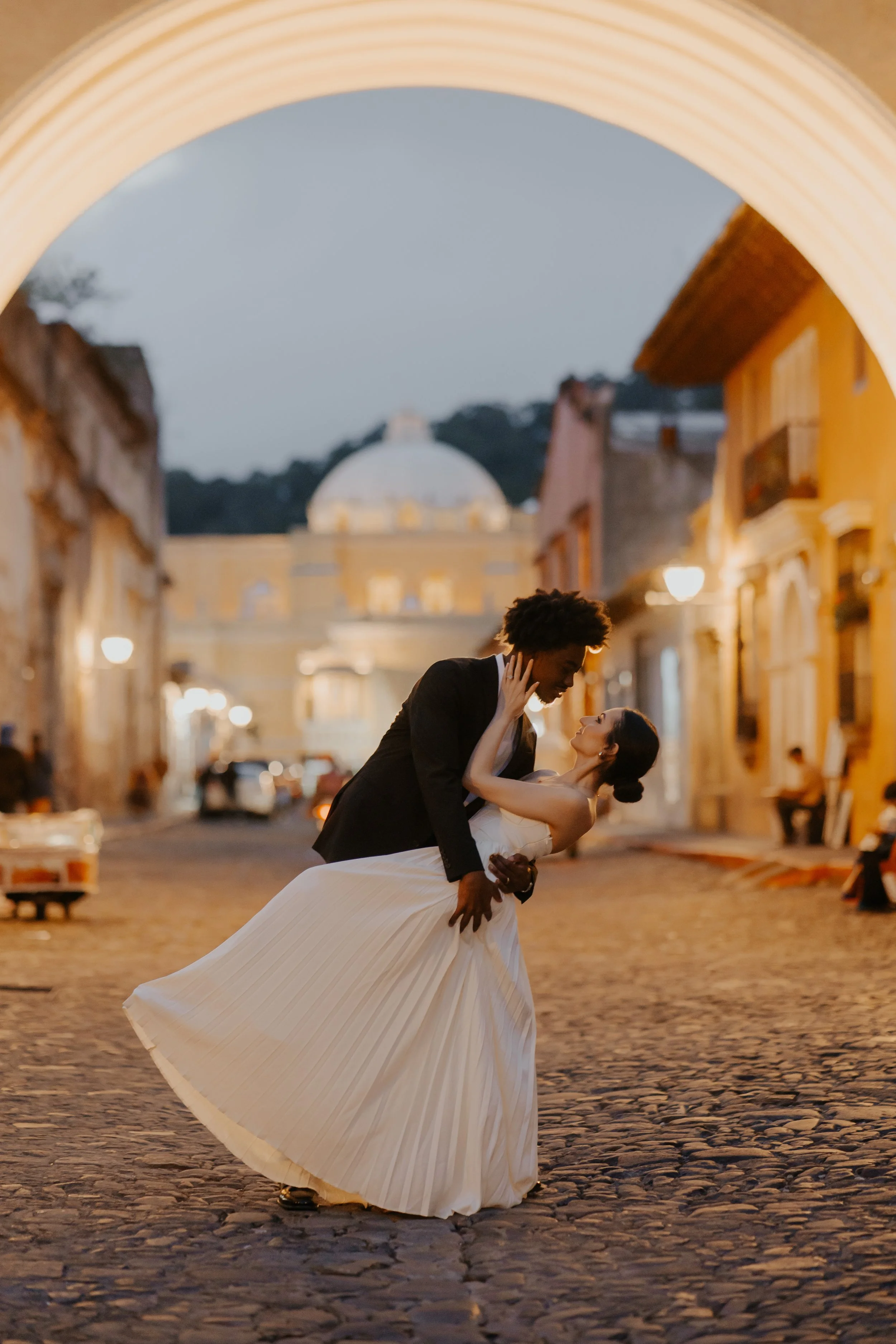 Pareja bailando en una calle empedrada al anochecer, con un fondo de edificios coloniales y un domo en la distancia.