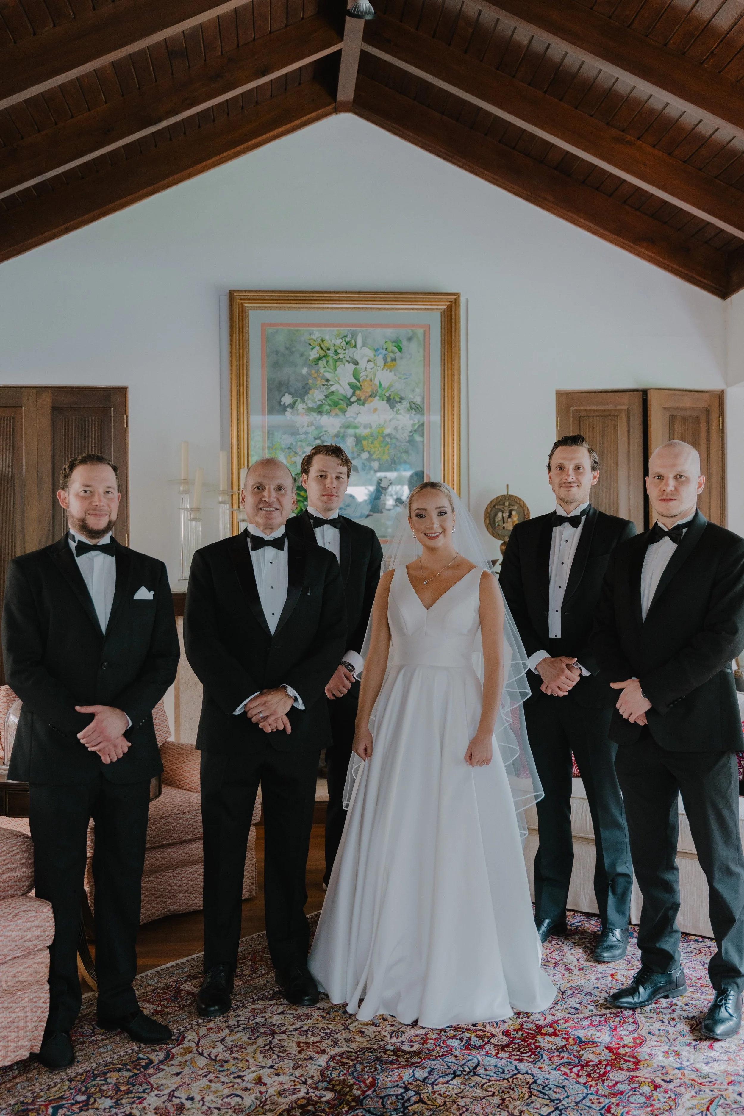 Un grupo de seis personas en una sala, incluyendo una novia con vestido blanco y cinco hombres con trajes de etiqueta, en un ambiente elegante con pintura y decoración en las paredes.
