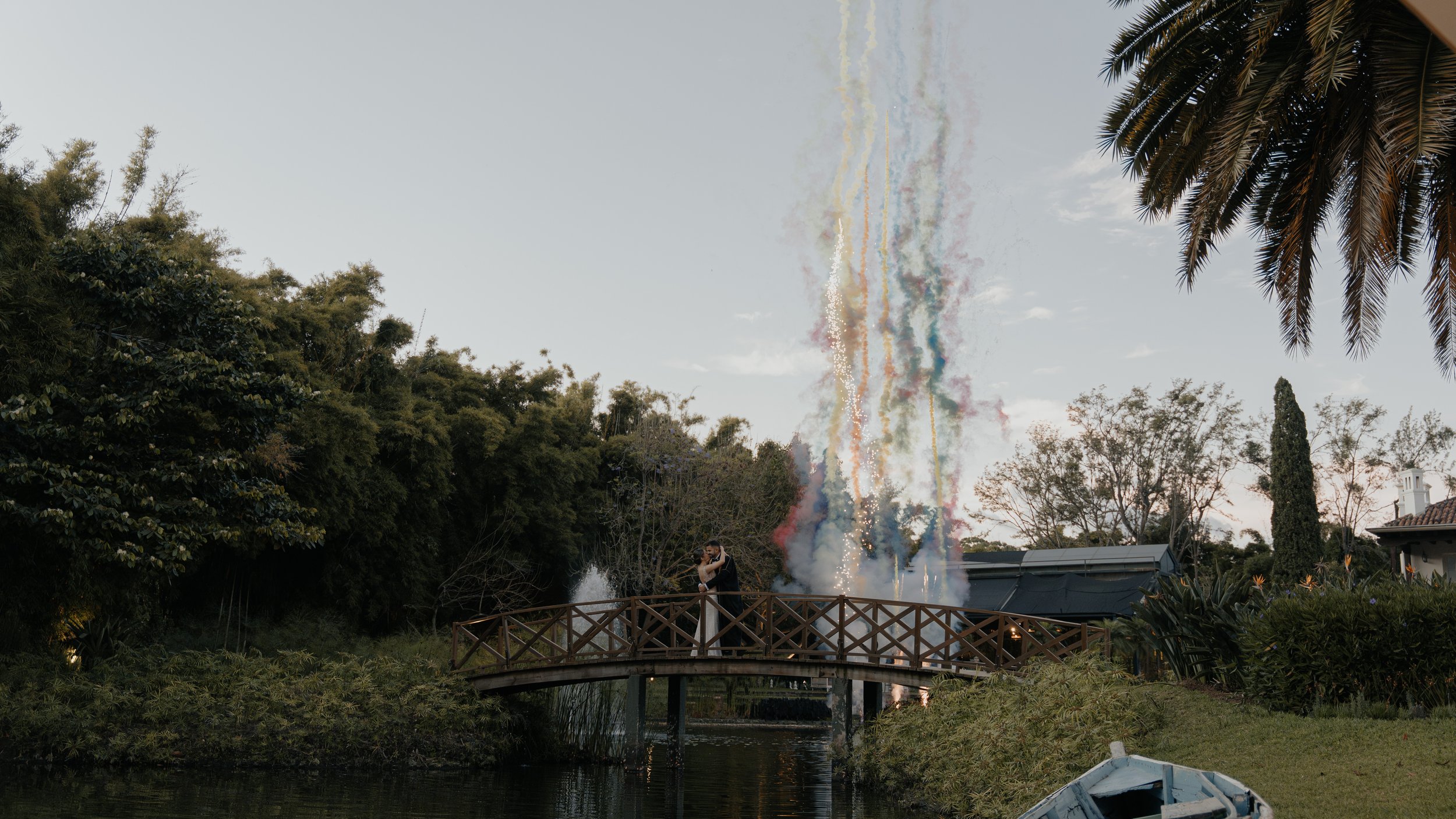 Una pareja de novios en un puente, besándose, con fuegos artificiales de fondo y árboles rodeando el área en un entorno al aire libre.