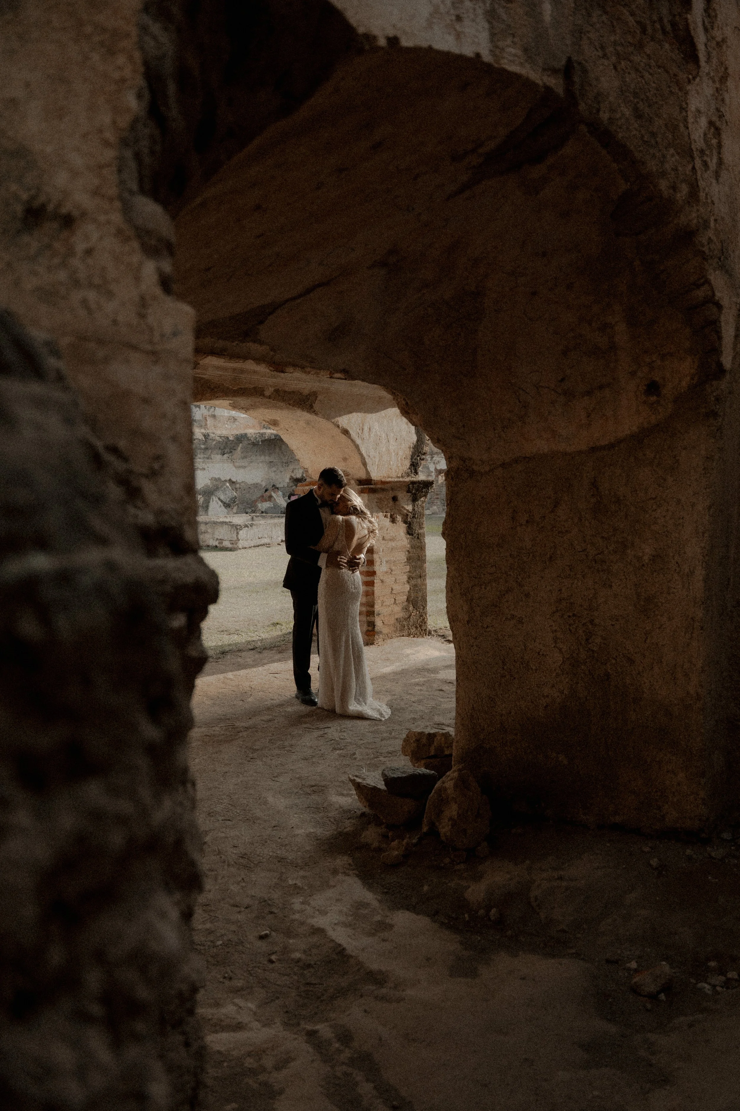 Pareja vestida de boda, abrazados en ruinas antiguas, vista a través de un arco de piedra