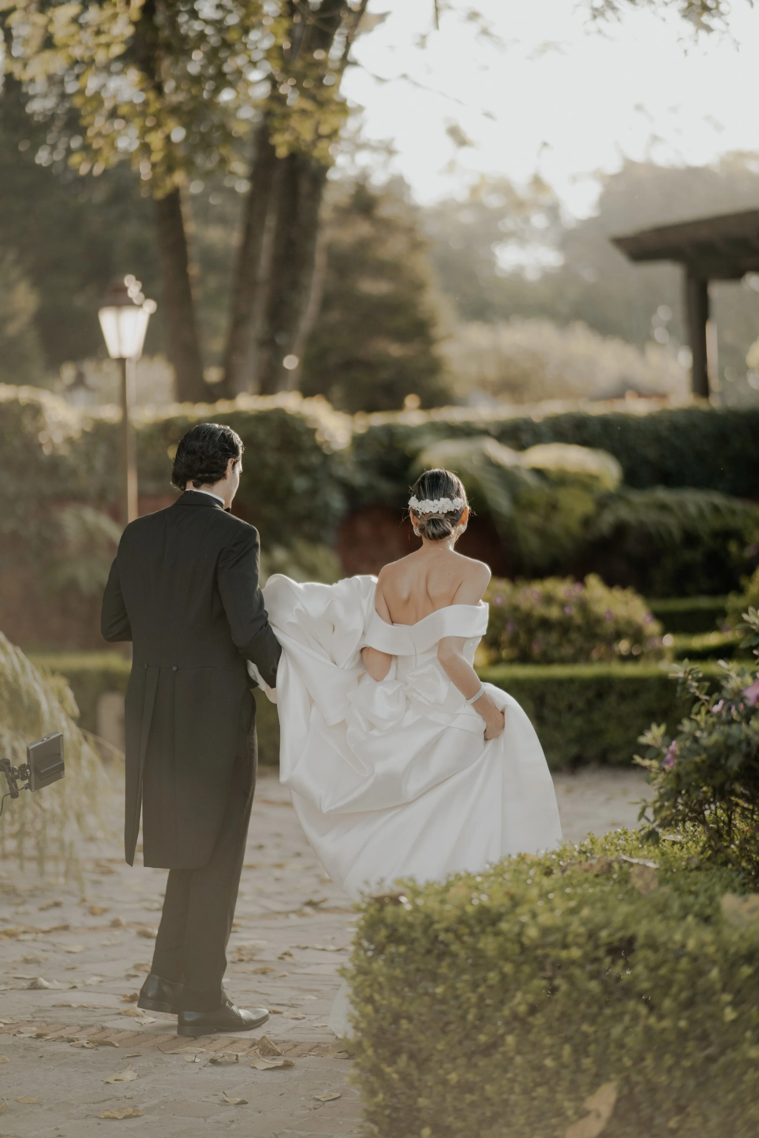 Pareja de novios caminando al aire libre en un jardín, la novia lleva vestido blanco y peinado con diadema, el novio usa traje negro.