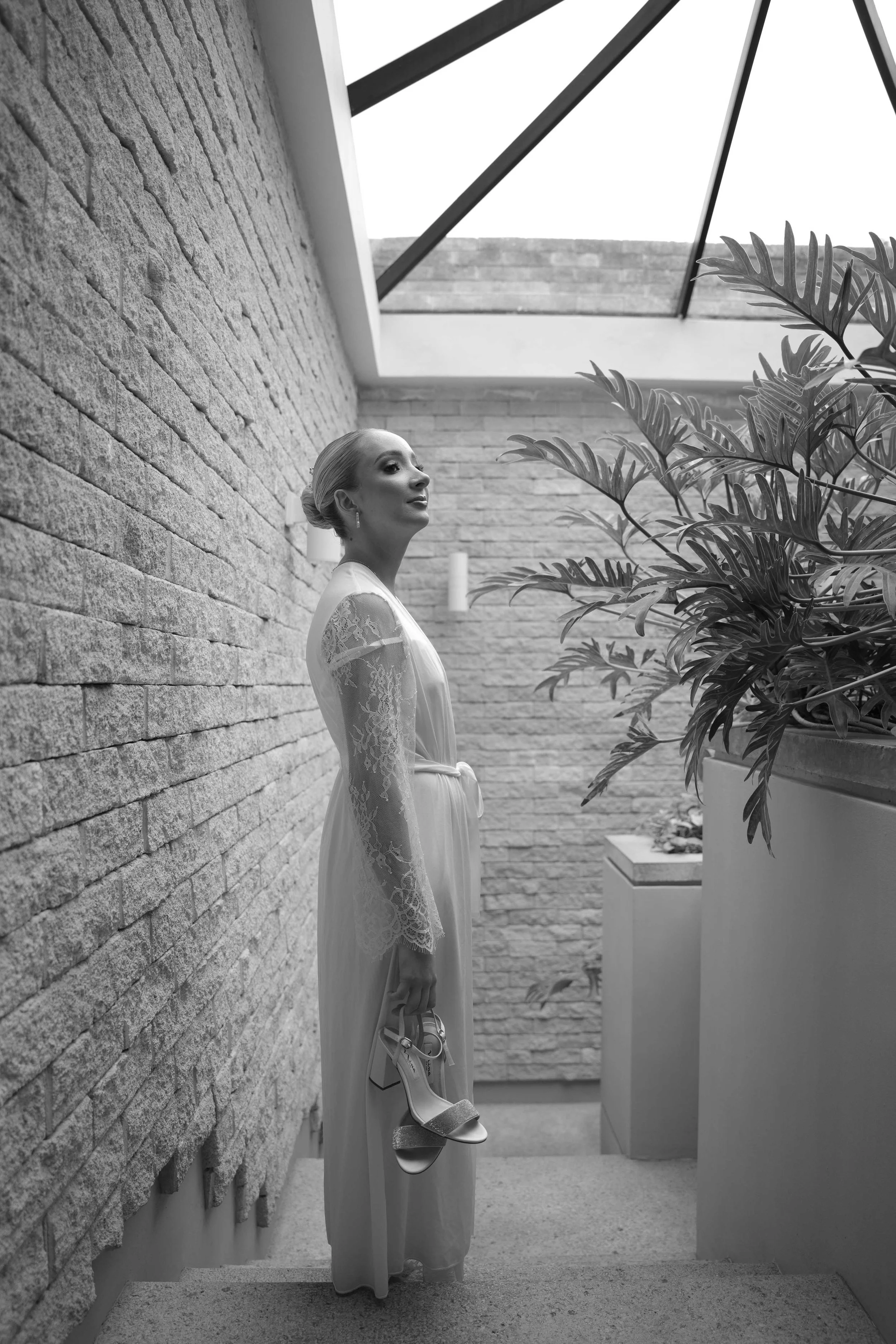 Mujer en vestido largo y con peinado elegante sosteniendo unos tacones en un espacio con paredes de piedra y plantas, en blanco y negro.