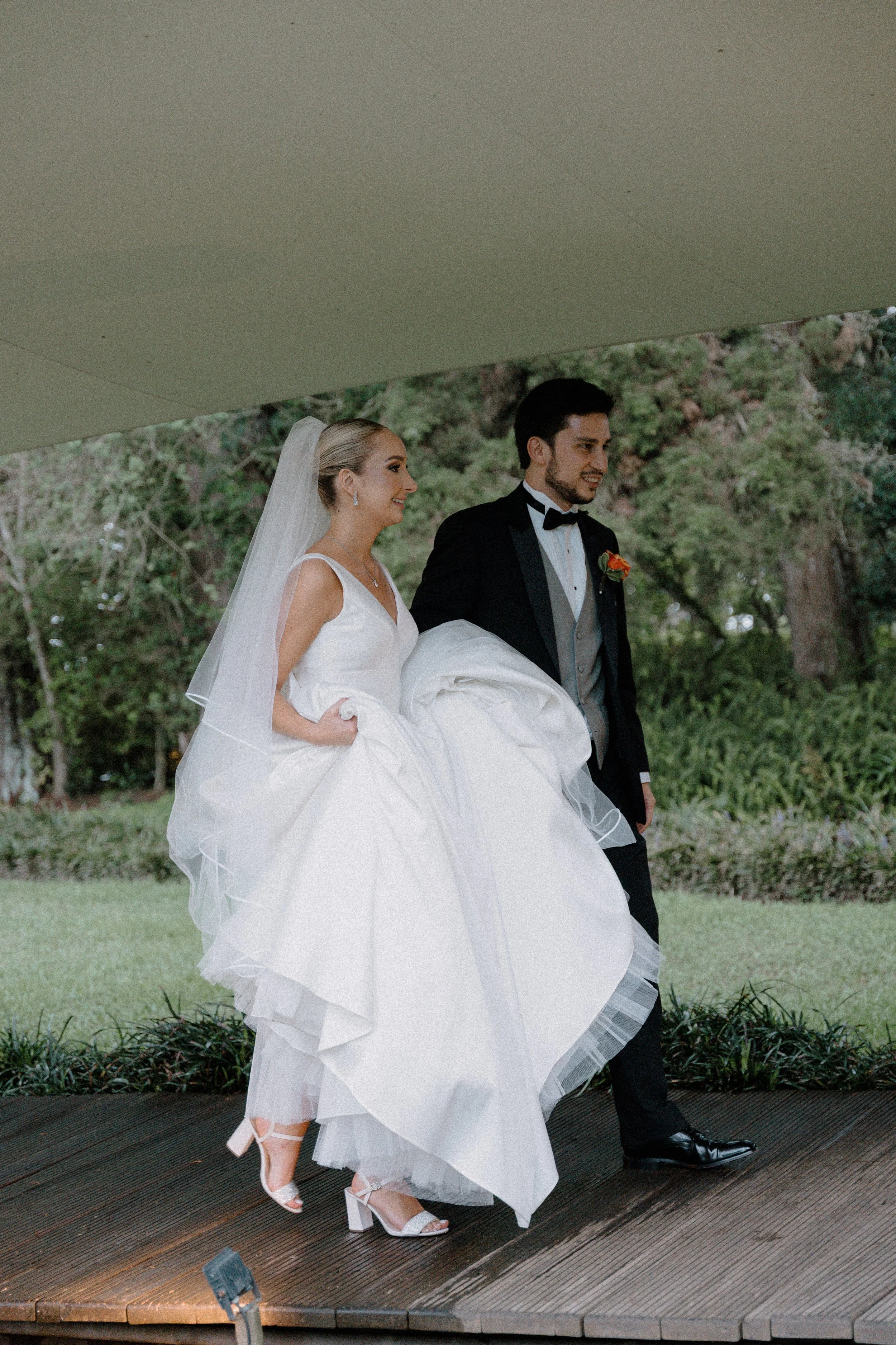 Pareja de novios en vestido de novia y traje de esmoquin caminando sobre un escenario exterior, con fondo de árboles y vegetación.