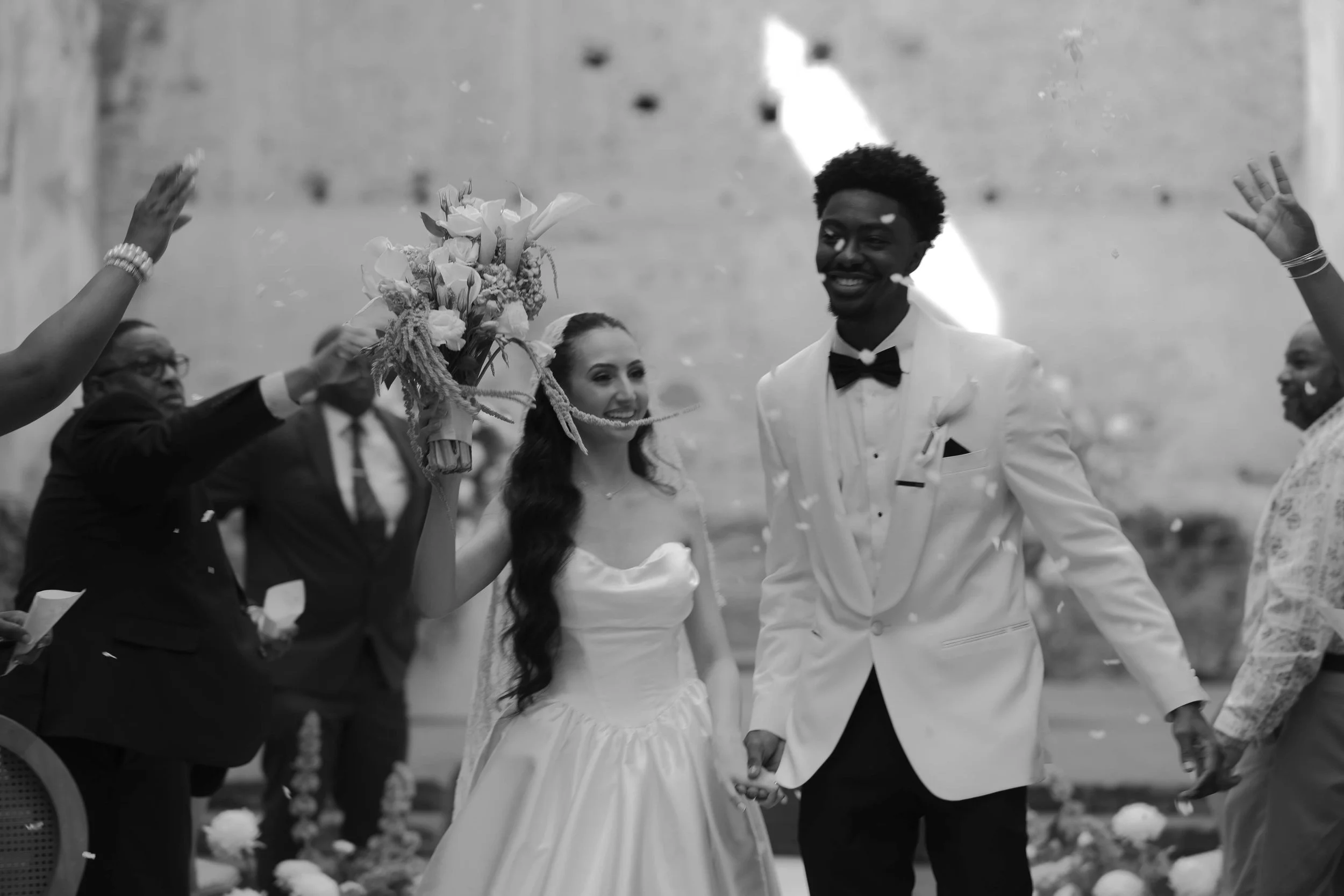 Una pareja de novios en una ceremonia de boda, ella sostiene un ramo de flores y se están dando la mano, rodeados de invitados en un lugar abierto.
