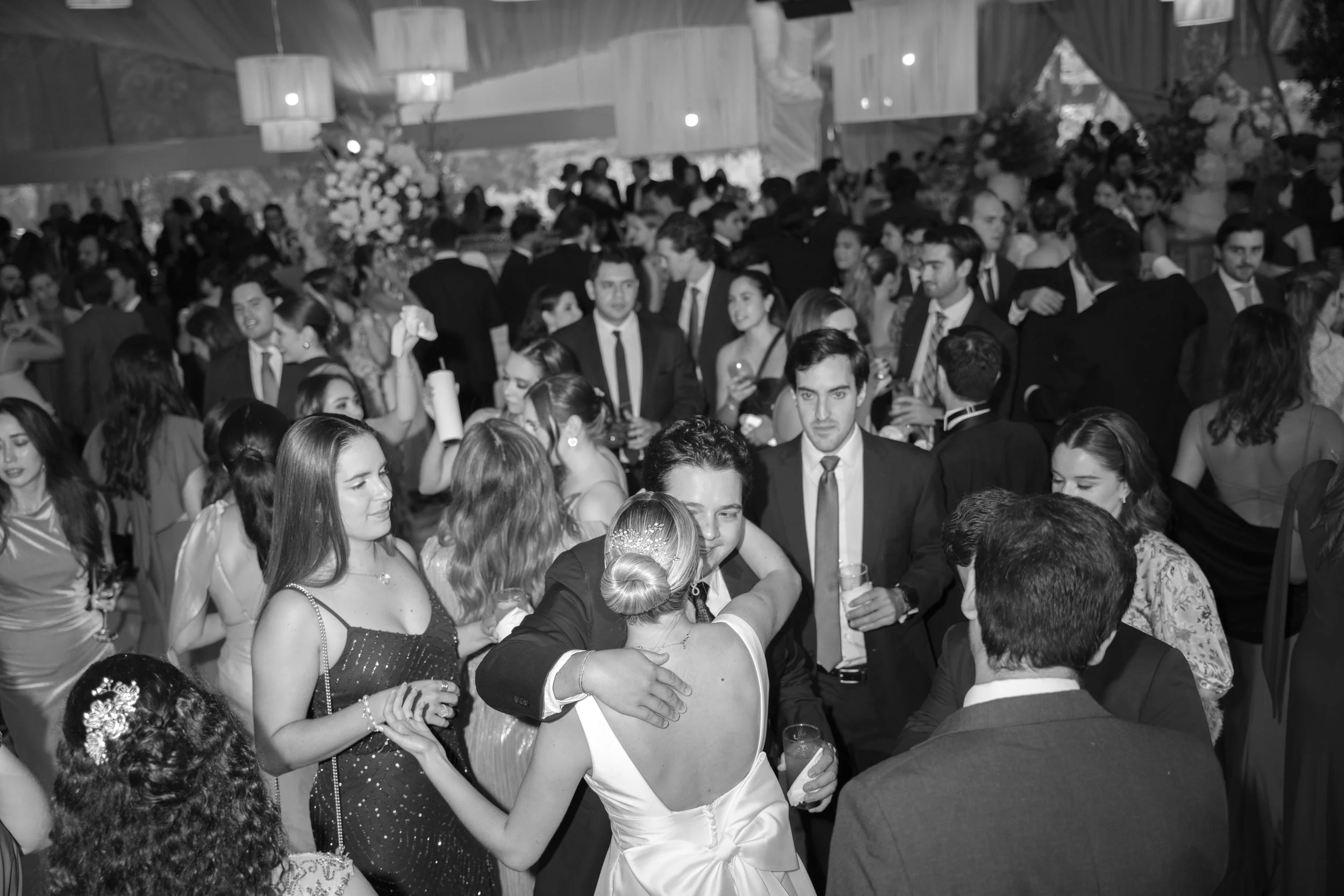 Pareja bailando en una celebración formal con muchas personas en el fondo, ambiente elegante con decoraciones florales y luces.
