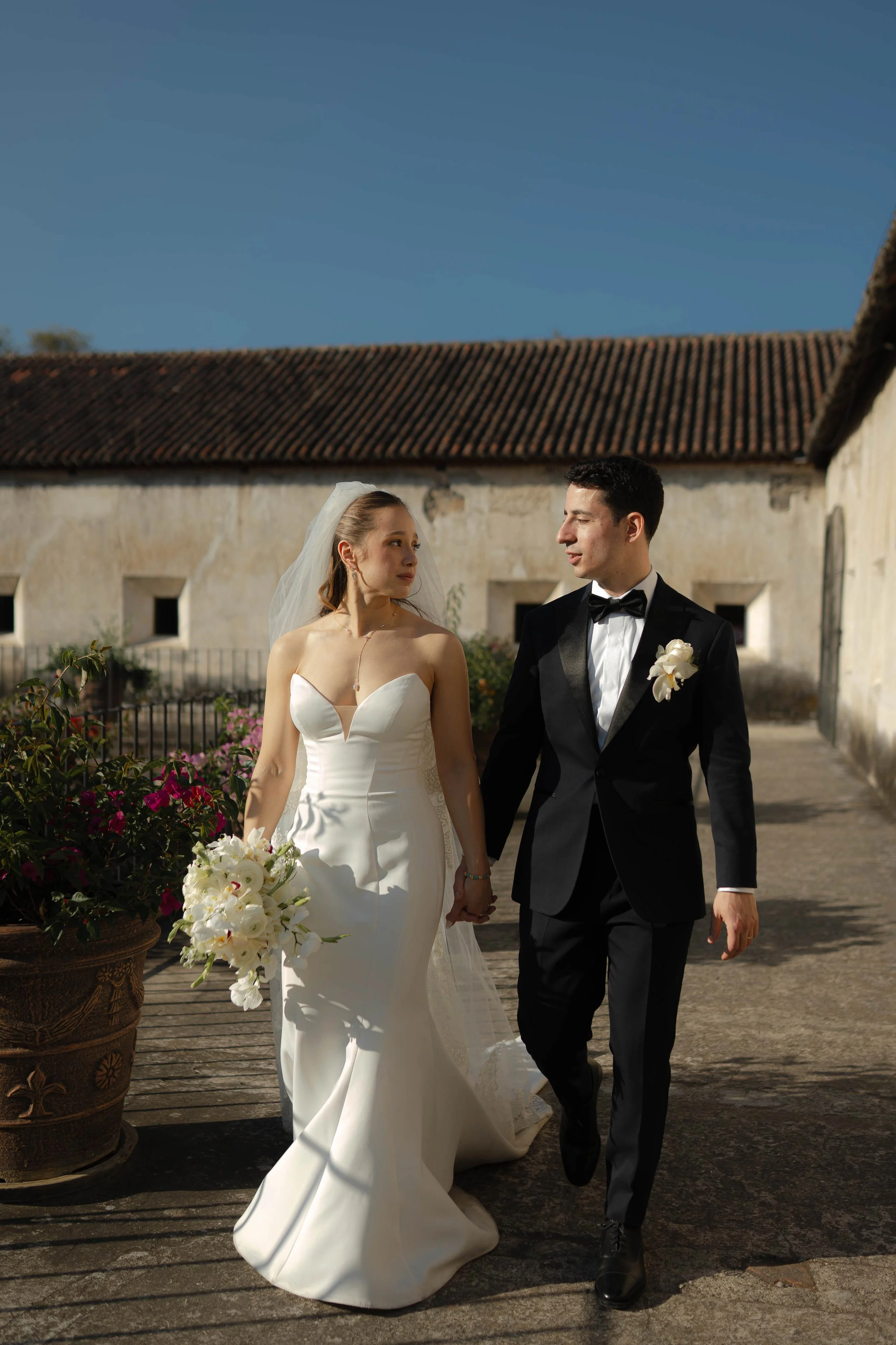 Pareja de novios caminando juntos en un escenario al aire libre, con la novia sosteniendo un ramo de flores blancas y ambos vestidos de manera formal para una boda.