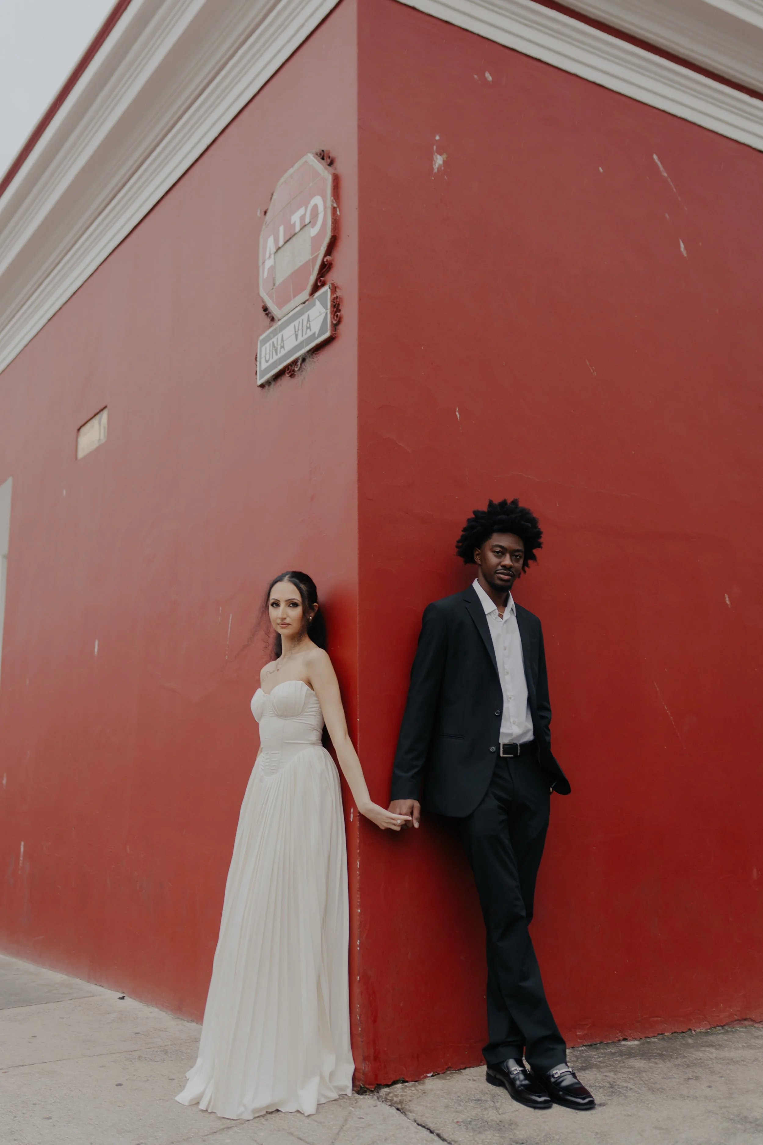 Una pareja vestida formalmente, sosteniéndose de las manos, apoyados en una esquina de una pared roja en una calle de ciudad.