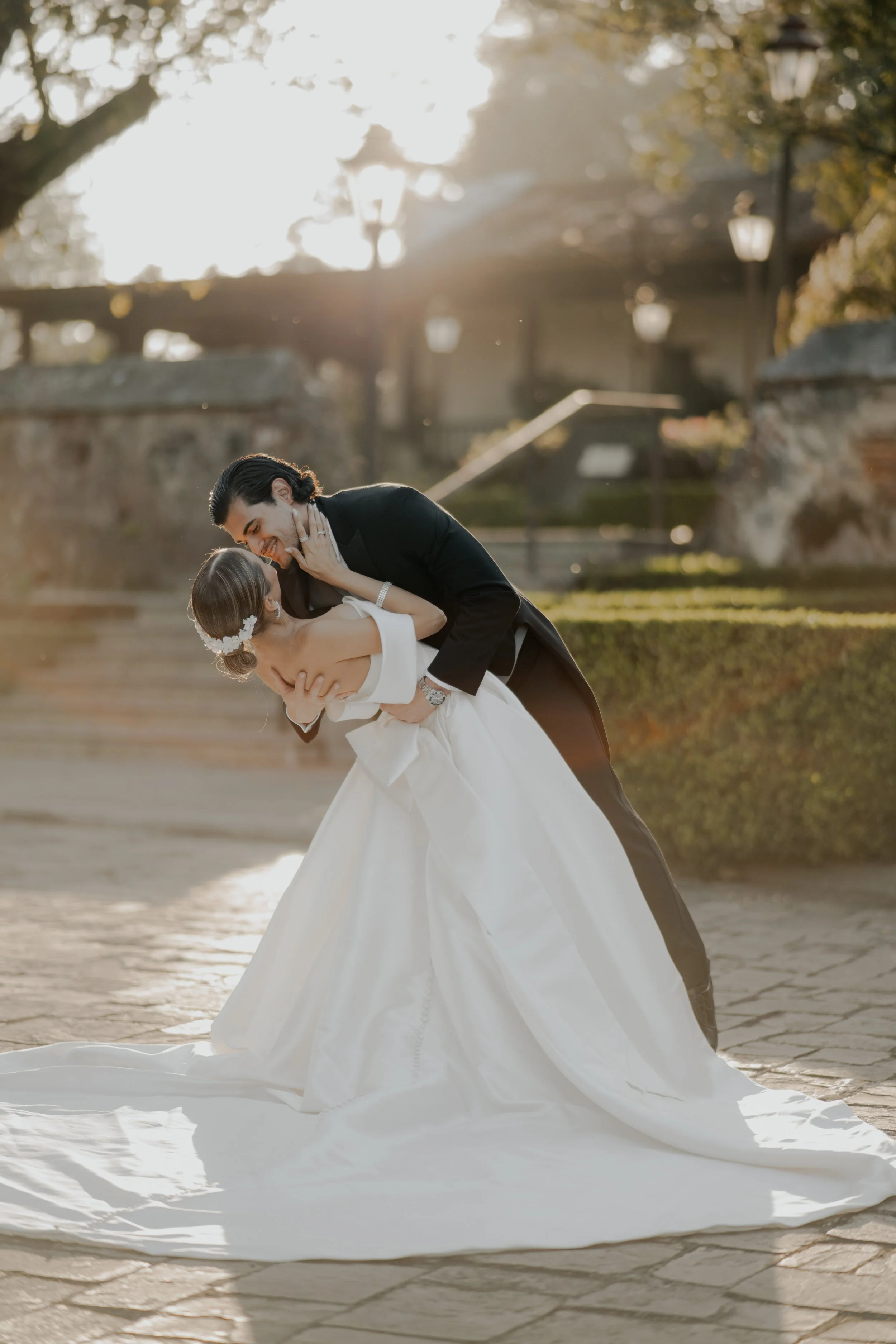 Una pareja de novios en su boda, el hombre la sostiene y la besa mientras ella sonríe, en un entorno al aire libre con sol de fondo y árboles.