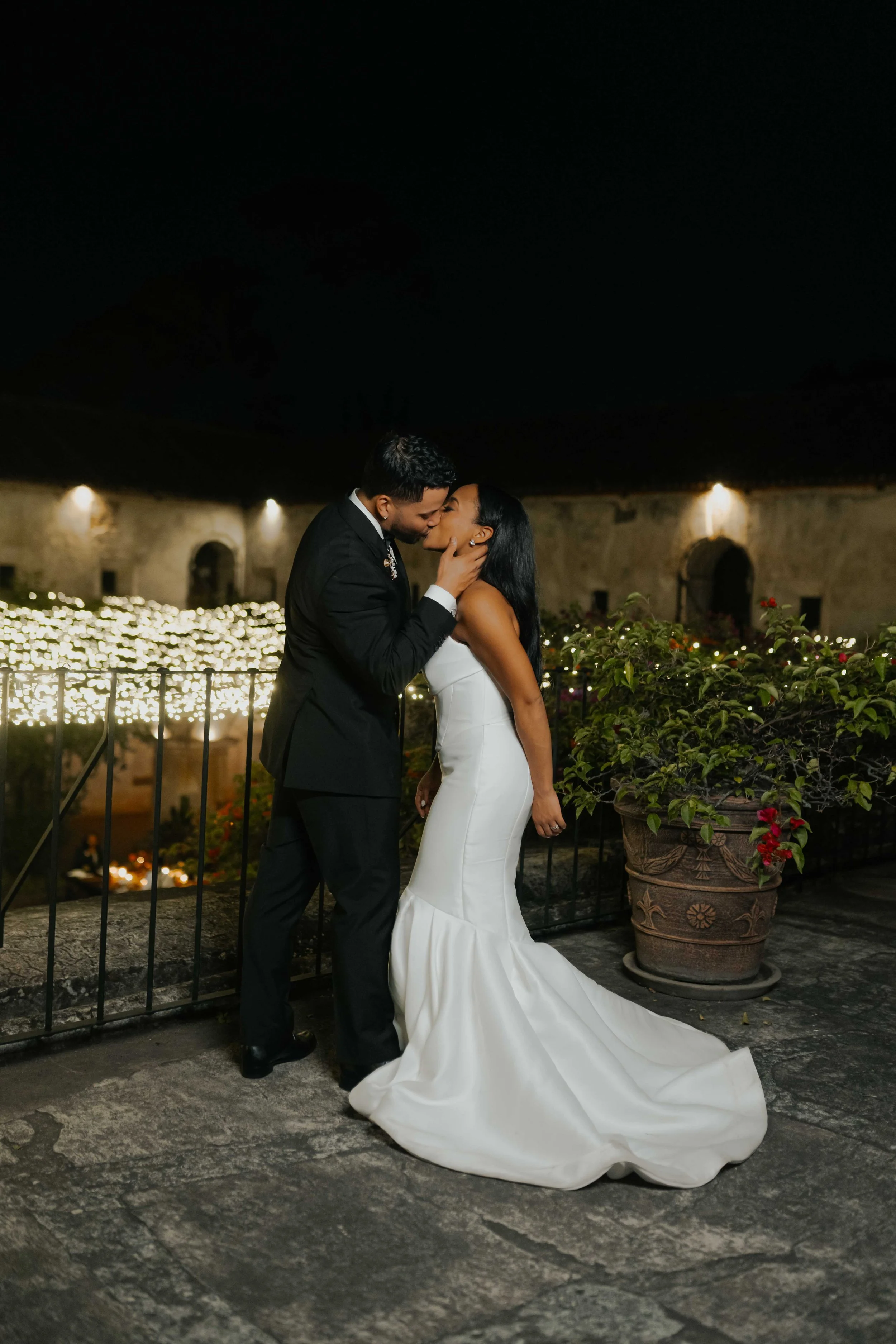 Pareja de novios besándose en una ceremonia de bodas por la noche, con luces y plantas de fondo.