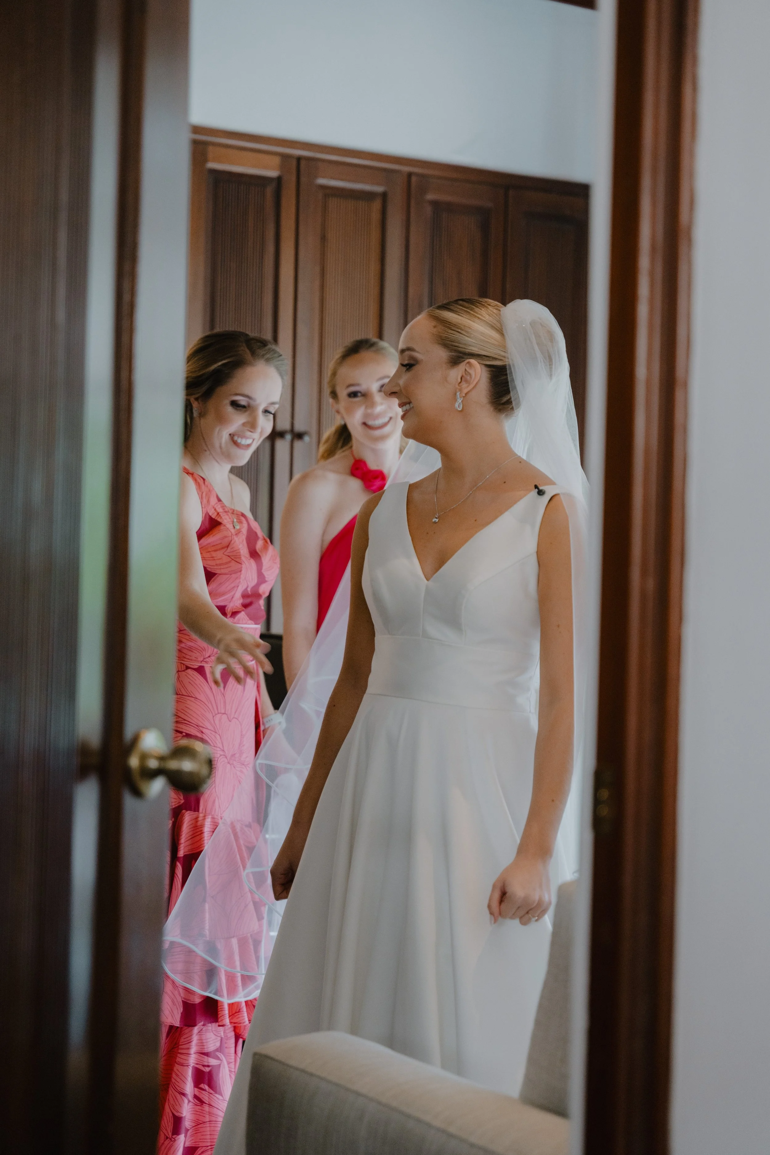 Mujer con vestido de novia sonriendo mientras otras mujer le ayudan con su vestido en una habitación