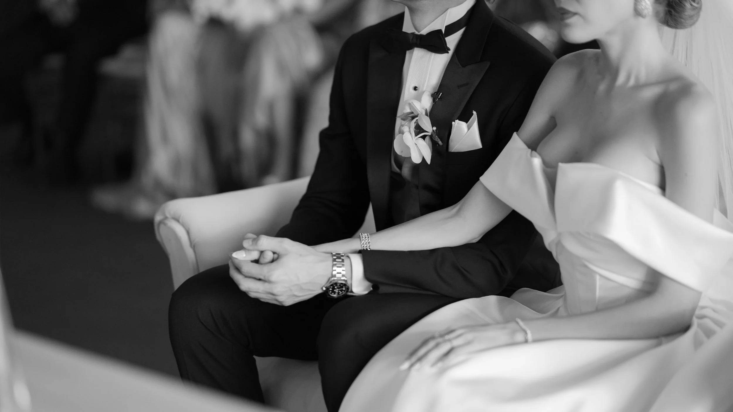 Pareja de novios en una ceremonia de bodas, tomados de la mano, sentados en un sillón, en un ambiente formal y elegante, en una foto en blanco y negro.