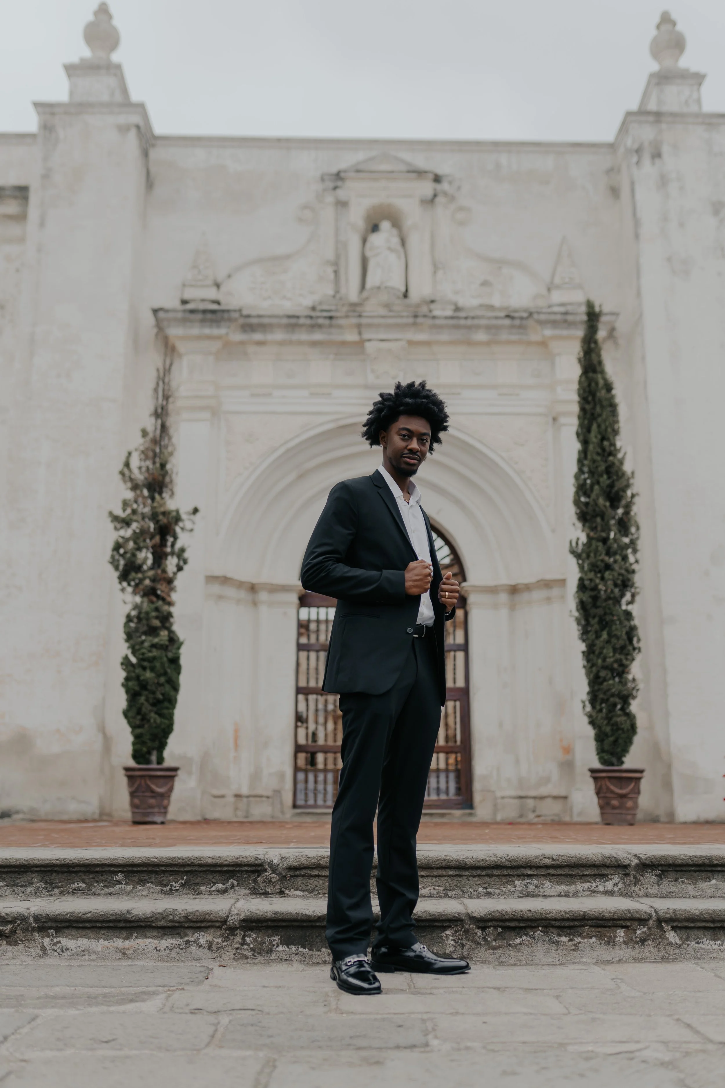 Hombre con traje negro de pie frente a un edificio colonial, con manos en las solapas, en una pose confiada.