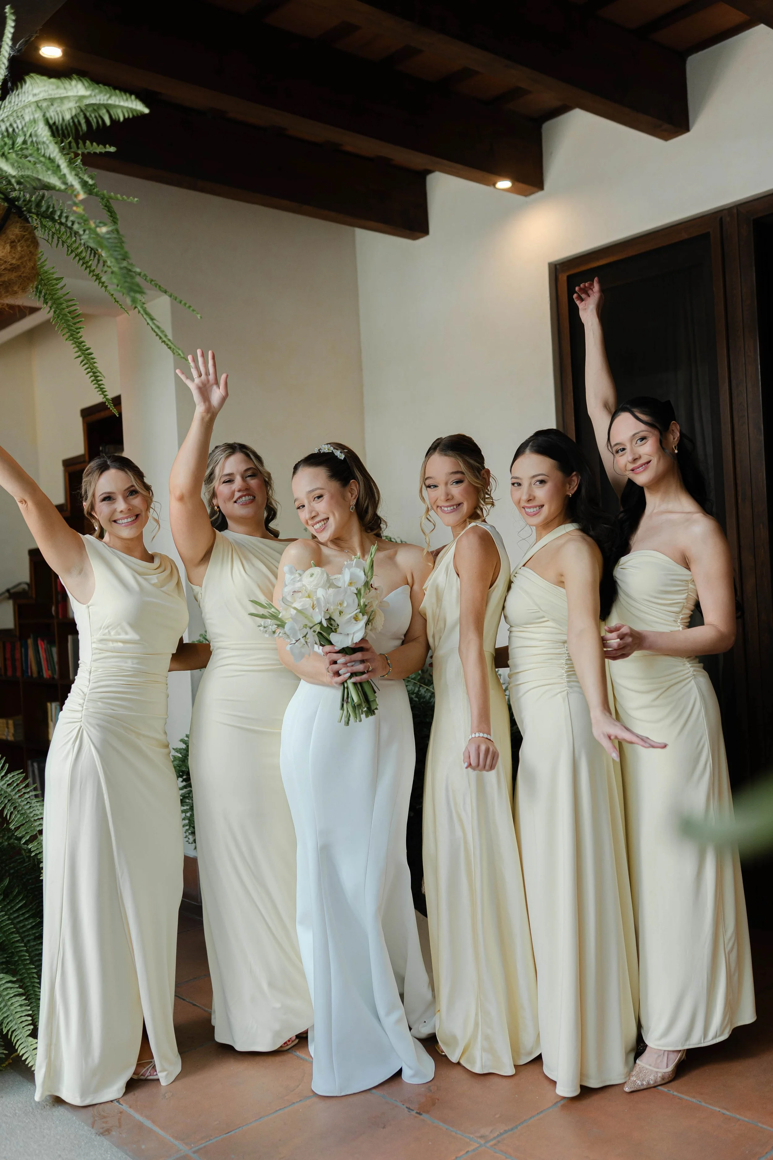 Grupo de mujeres en vestidos claros, celebrando una ocasión especial, con una de ellas sosteniendo un ramo de flores.