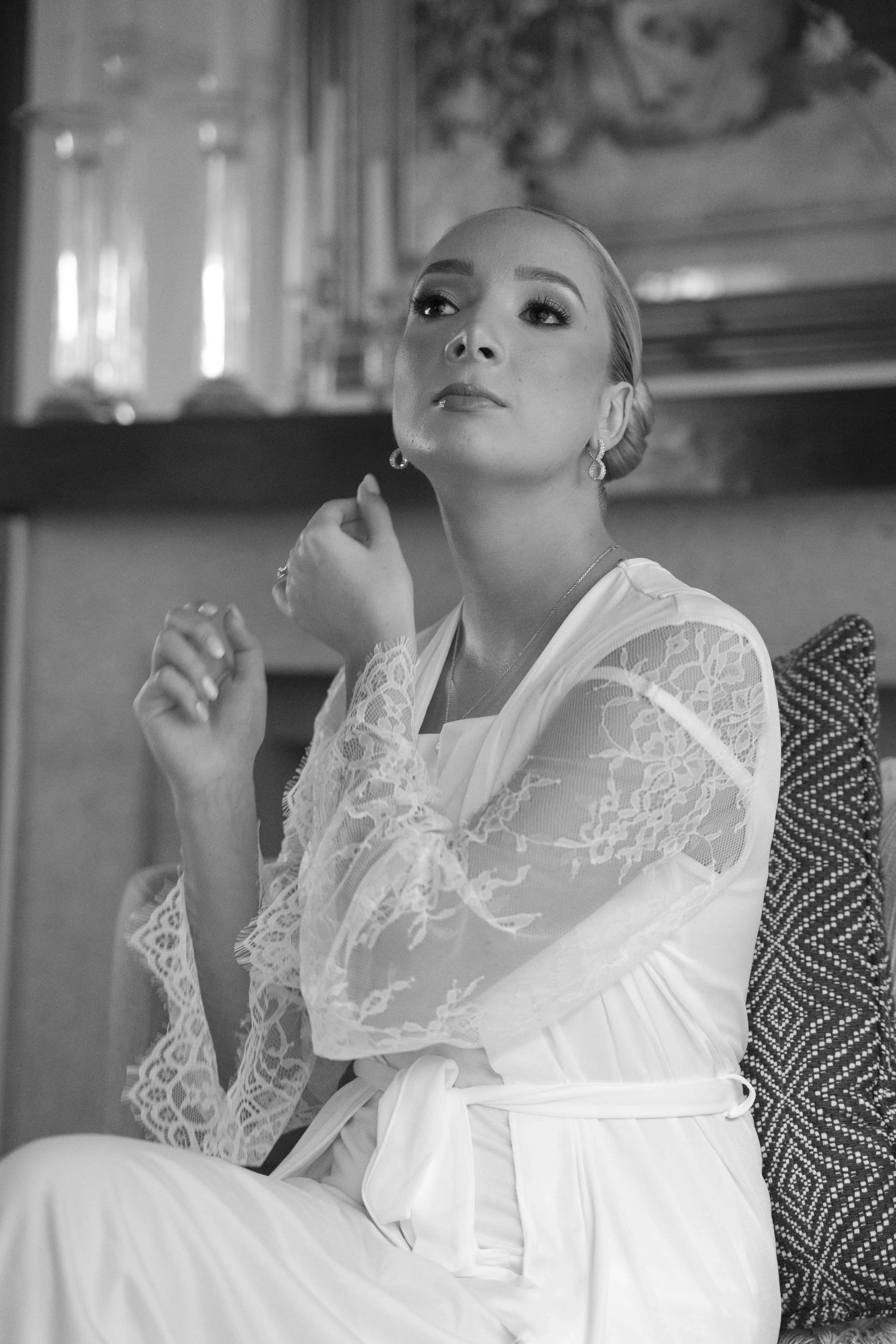 Retrato en blanco y negro de una mujer con cabello rubio, luciendo elegante con aretes y ropa de encaje, sentada en una silla con cojín decorativo, en un ambiente interior con decoración clásica y cuadros en la pared.